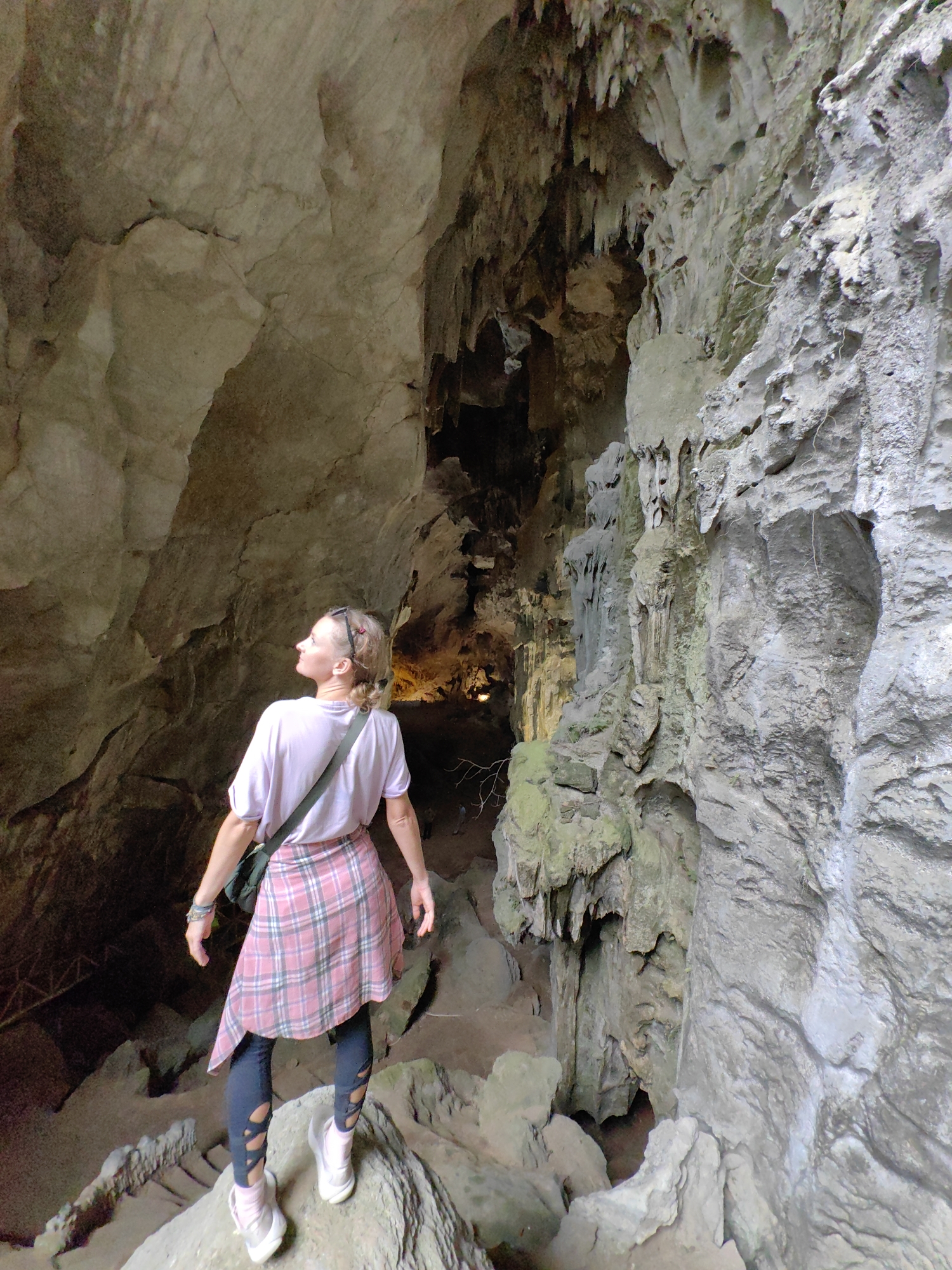 A woman stands in a cave, looking up and towards the interior. The cave is rocky, with formations of stone and possible stalactites. Light sources illuminate the deeper part of the cave. The woman is wearing a pink shirt, a plaid skirt tied around her waist, dark leggings with cutouts, and light-colored sneakers. She has a shoulder bag. The location is likely in or near Lạng Sơn, Vietnam.