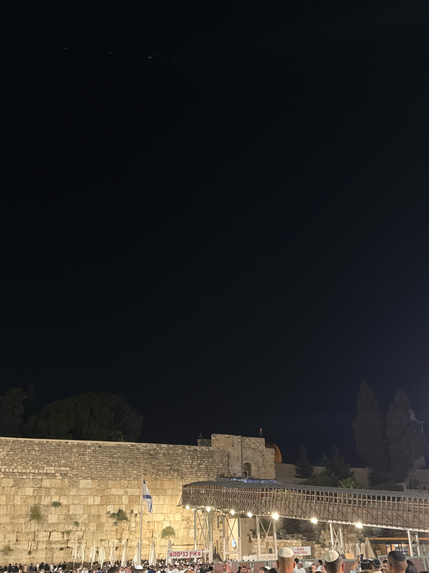 The image captures a nighttime scene at the Western Wall in Jerusalem, Israel. A large, ancient stone wall, likely the Western Wall itself, dominates the lower half of the frame, illuminated by artificial lights. In front of the wall, a crowd of people, many wearing white shirts and kippahs, are gathered. Some appear to be praying or observing. A walkway or bridge structure, made of wood and metal, is visible in the middle ground, spanning across the plaza. An Israeli flag is planted on a flagpole near the center of the image. In the background, silhouetted trees and parts of buildings, including a dome, are visible against the dark night sky. Signs with Hebrew lettering are present, adding to the cultural context of the location. The overall impression is one of a busy religious site at night, with a significant number of people present.