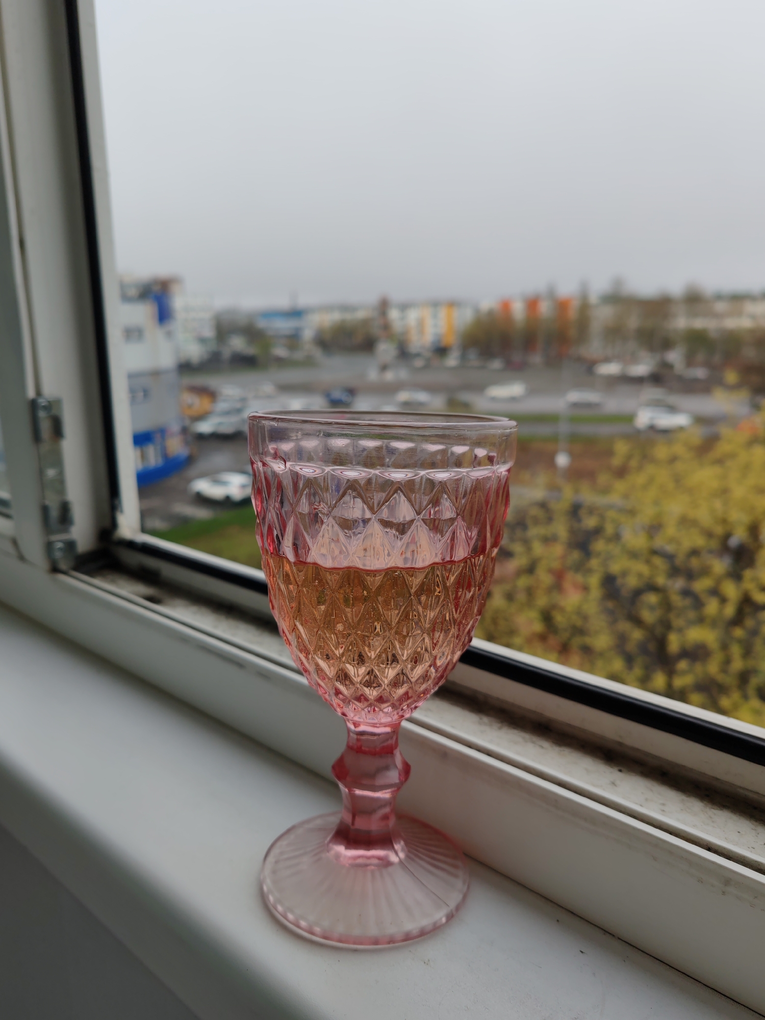 A pink glass goblet, about half-filled with a light-colored liquid, stands on a white windowsill. The window is open, showing a blurred cityscape. Cars are visible on the roads. Buildings with colored facades (blue, white, orange) are in the background. Some yellow foliage is present. The scene appears to be in Petropavlovsk-Kamchatsky, Russia. The weather is overcast. The window frame is white with some visible dirt and discoloration.