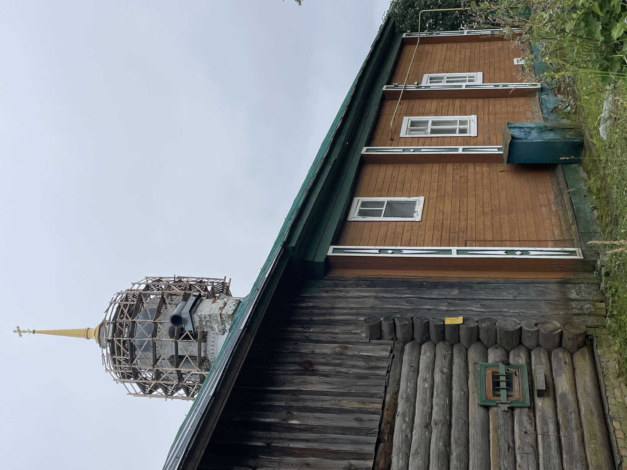 In the city of Кстинино, Russia, a wooden log building is visible in the foreground, its logs weathered and gray. To the right, a section of the building is covered in horizontal wooden planks, painted a warm brown. This section features several white-framed windows with green trim. A green, weathered metal box sits on the foundation near the right side of the brown-planked section.
Behind the buildings, a church dome under scaffolding is prominent. The dome is golden and topped with a cross. The scaffolding is constructed of light-colored wood. A brick chimney protrudes from the roof of the log structure, near the base of the dome. The roof of the log building is covered in dark material, possibly asphalt shingles, and has green trim along the edge. The sky is overcast and a pale gray. Wild vegetation, including tall grasses and flowering plants, grows around the base of the buildings.