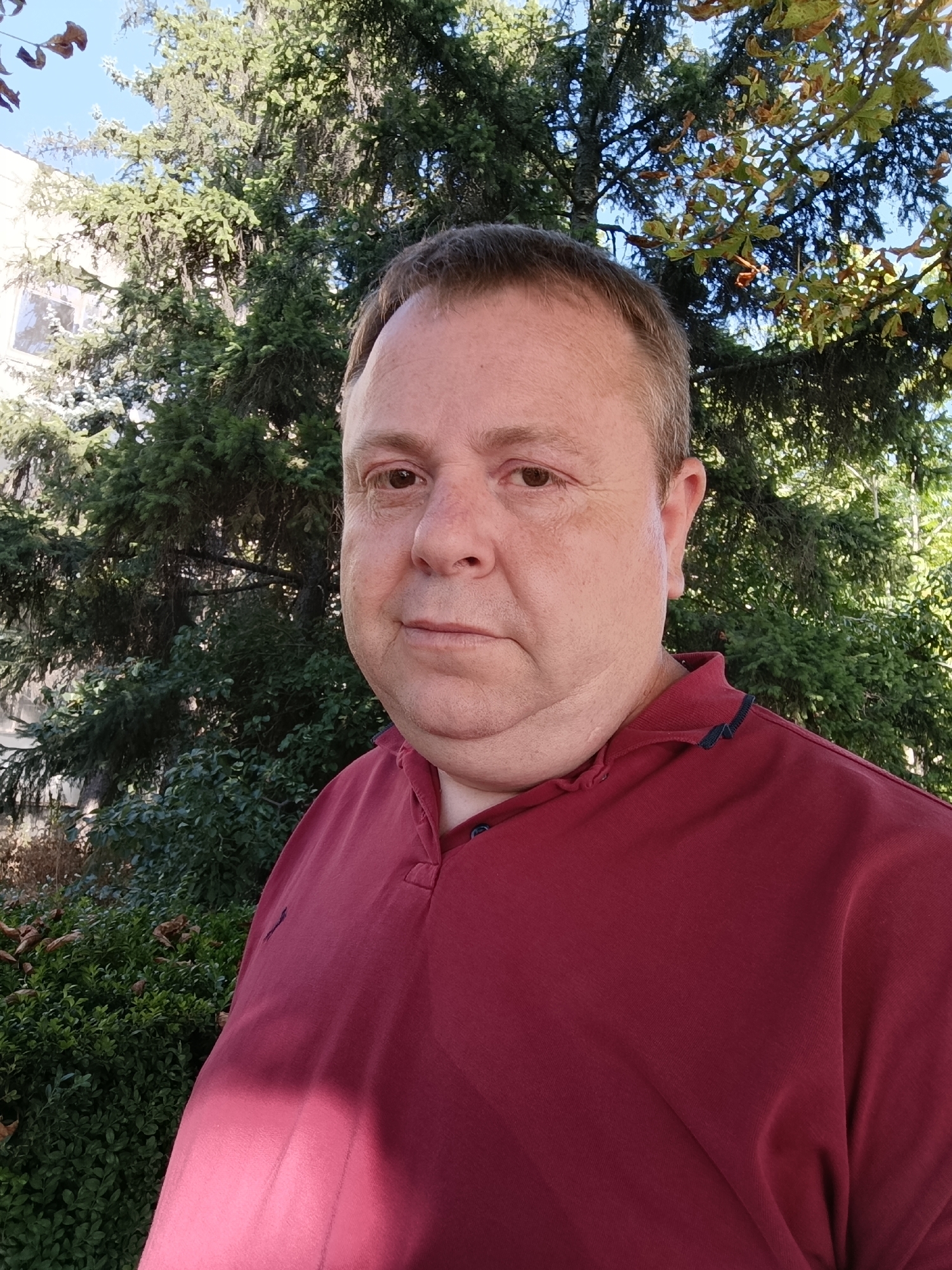 A medium shot shows a man in his late 40s or early 50s standing outside, with trees and a building in the background. The man is facing the camera, with his head tilted slightly to his right. He has a fair complexion, light brown hair, and brown eyes. He is wearing a maroon polo shirt with dark blue trim around the collar. The background consists of a large evergreen tree, some smaller green bushes, and a light-colored building. The lighting suggests it is daytime. The image appears to have been taken in the city of Dubăsari, Moldova.