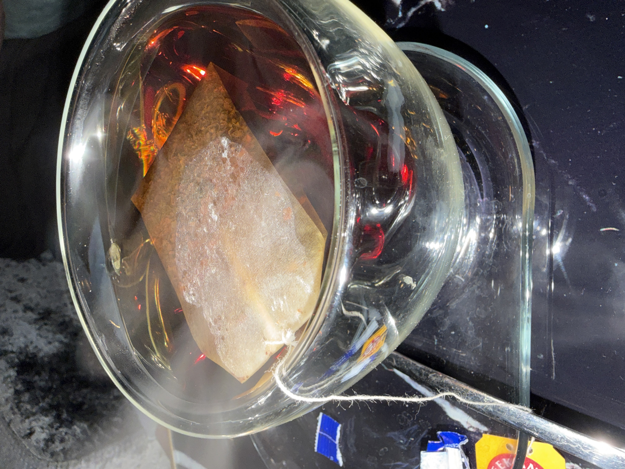 In Nagold, Germany, a close-up shot focuses on a clear glass bowl filled with hot, dark tea. A tea bag, made of a light-colored, porous material, is immersed in the tea, with its string dangling over the side of the bowl. Wisps of steam rise from the hot liquid, indicating its temperature. The bowl rests on a black, reflective surface, possibly a tray or table, which shows faint, colorful reflections of light. Small, colorful, abstract-shaped elements are visible on the black surface near the base of the bowl, possibly decorative elements or debris. The lighting is somewhat dim, creating dramatic highlights and shadows, particularly on the glass bowl and the surface below. The overall scene evokes a sense of warmth and relaxation.