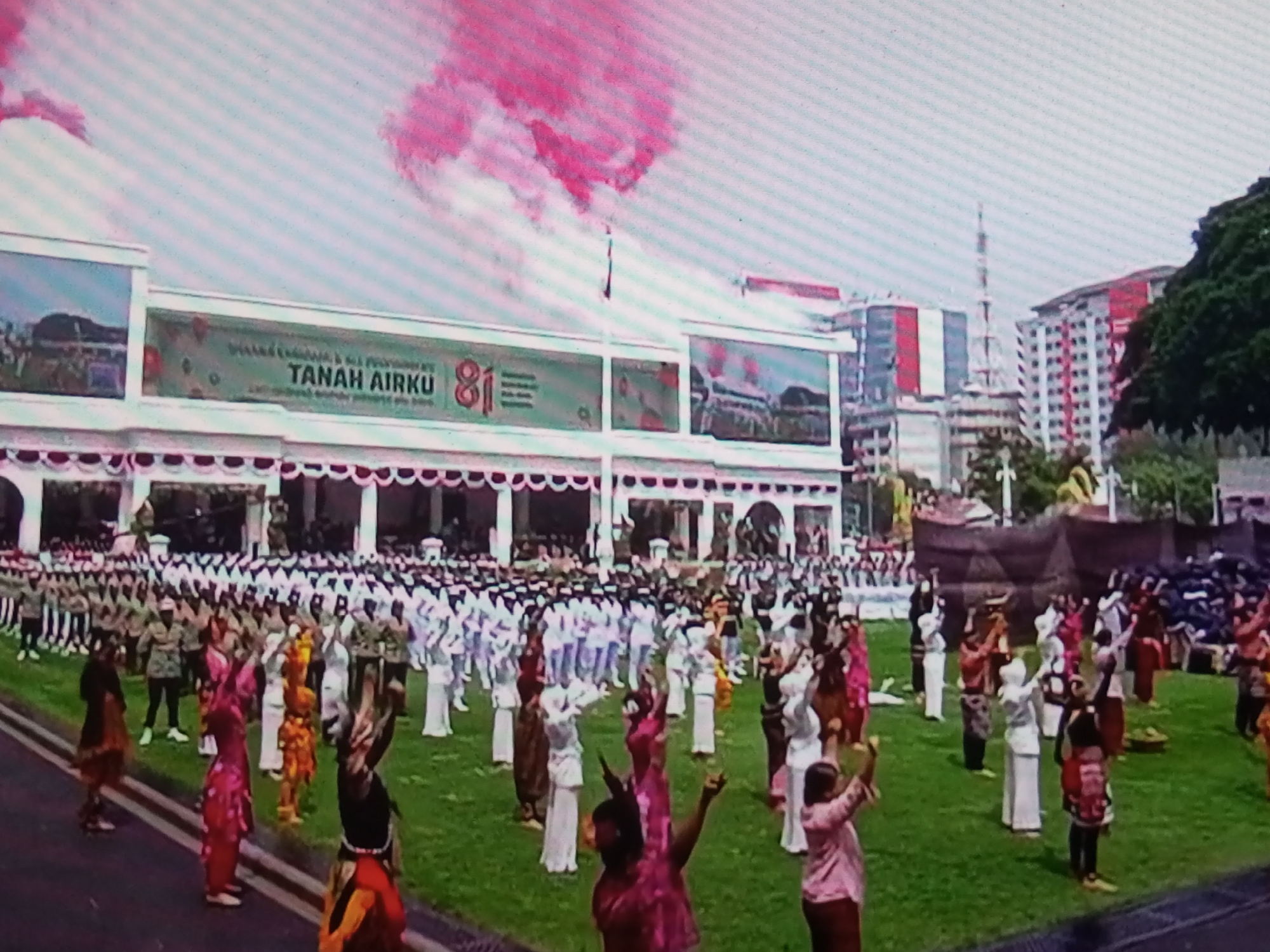 This is a wide shot of an outdoor event, likely a parade or festival, taking place in the city of Banyudono, Indonesia. A large white building with a banner that reads "TANAH AIRKU" (My Homeland) and the number "81" is in the background, along with some trees and modern buildings. A large crowd of people, dressed in various uniforms and traditional attire, are gathered on a grassy field. Some are marching, while others are performing a choreographed dance. There is a plume of pink smoke rising into the sky, suggesting fireworks or a smoke bomb. The event appears to be a celebration of some kind, possibly a national holiday or anniversary. The overall atmosphere is festive and patriotic.