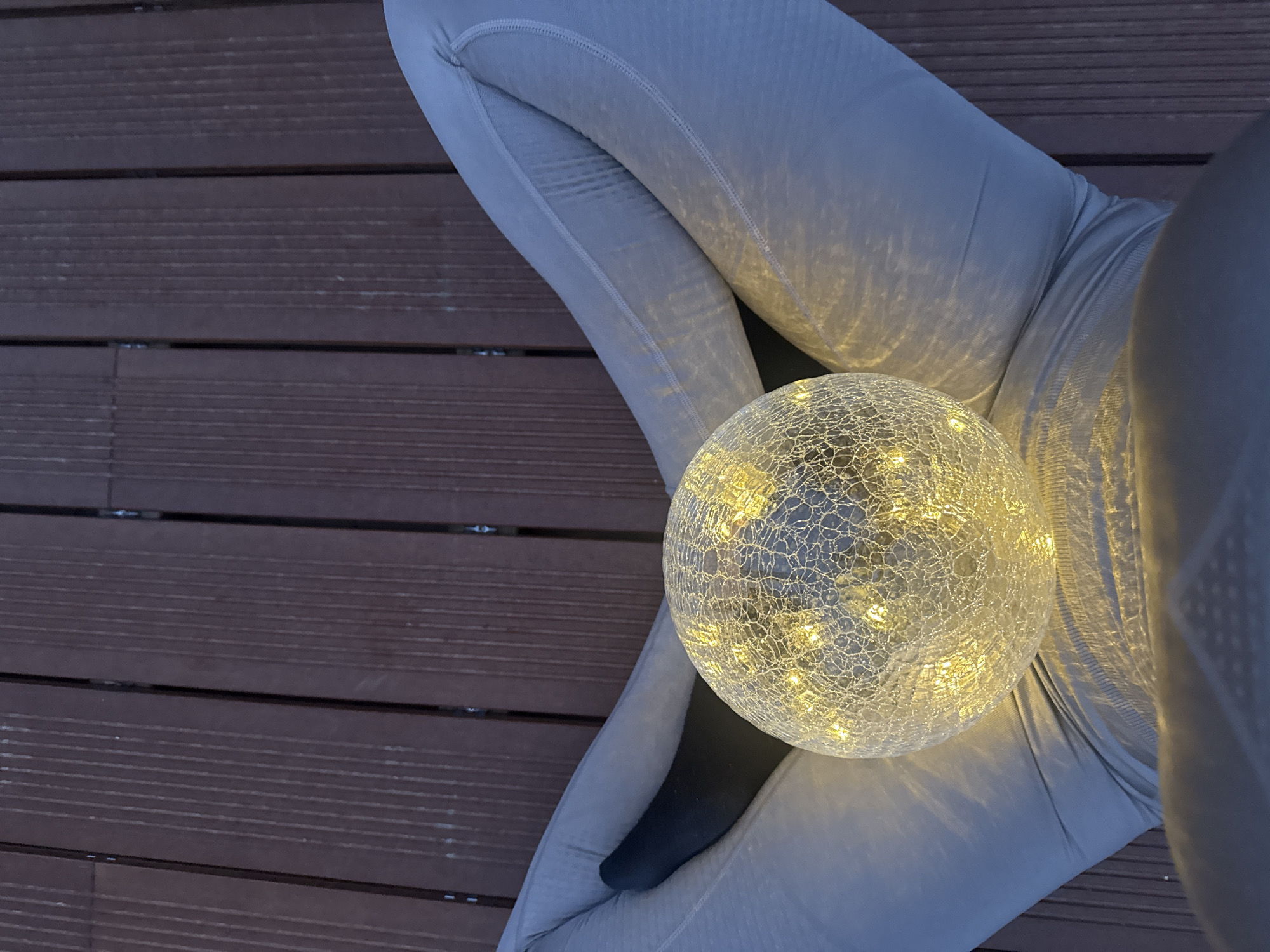 A top-down shot shows a person wearing light gray leggings sitting on a dark brown wooden deck. A decorative light-up sphere with a cracked-glass pattern rests in their lap. A small portion of a black sock is visible. The environment is outdoor, potentially a balcony or patio. The location is not discernible from the image.