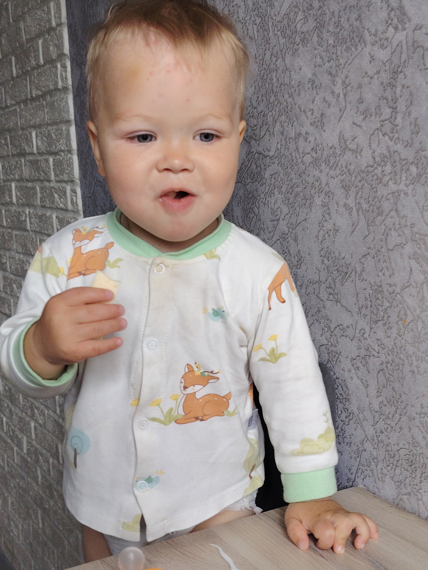 A fair-skinned toddler with light blonde hair stands next to a light brown table, with a light gray brick wall and textured gray wall in the background. The child, who appears to be eating, is wearing a white long-sleeved pajama top with a green trim, featuring images of deer and plant life. The toddler holds an object in his right hand near his mouth. The location is presumed to be an indoor space in Chaikovsky, Russia, based on provided information.