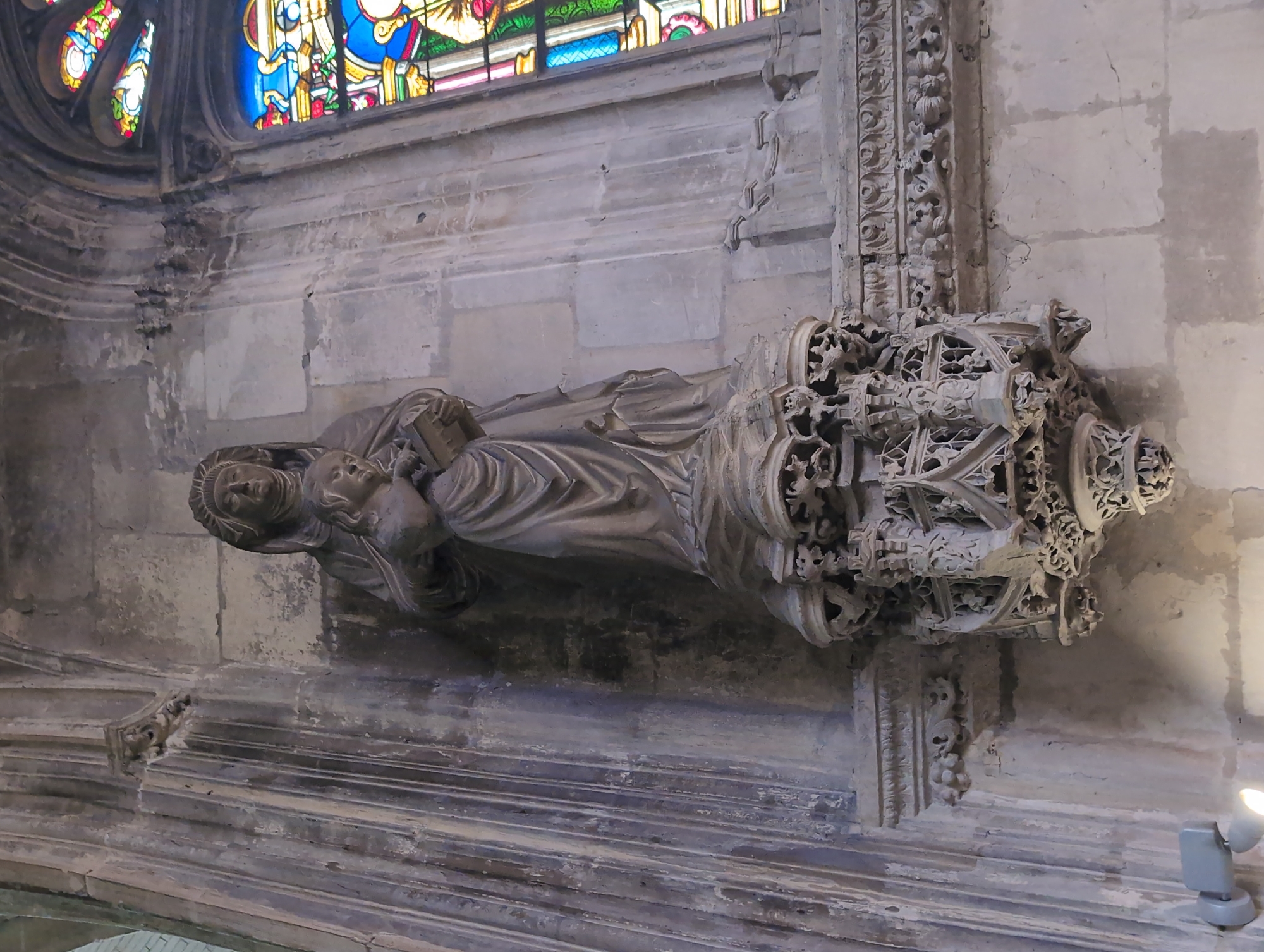 A full shot captures a weathered stone sculpture of the Virgin Mary holding the infant Jesus, mounted on a column within a dimly lit stone interior, likely a church or cathedral. The sculpture, in a Gothic style, depicts Mary with a serene expression, her face covered by a veil, and Jesus as a young child with a book in his hands. The column is elaborately carved with ornate details, forming a tiered, openwork pedestal that supports the figures. The background features a stained-glass window with vibrant blues, reds, and yellows, and sections of rough-hewn stone walls. The lighting is low, with a visible light fixture mounted to the wall near the base of the column. This sculpture is located in the Church of Saint-Jacques in Dieppe, France.
