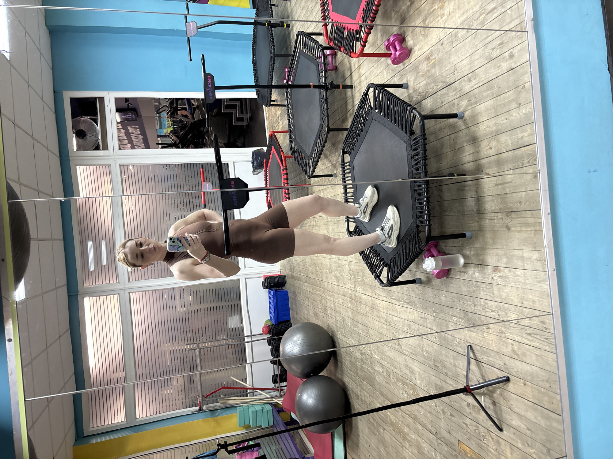The image captures a young woman taking a mirror selfie in a fitness studio in Saint Petersburg, Russia. The woman, wearing a form-fitting brown athletic one-piece and white sneakers, stands on a black mini-trampoline, holding a smartphone with a floral case up to capture her reflection. Her blonde hair is pulled back, and she has a neutral, slightly pouting expression.
The fitness studio features light wooden floorboards and a light blue wall. Several mini-trampolines, some black and others with red accents, are arranged across the floor. Pink dumbbells are visible near the trampolines, and a clear water bottle is beside the woman's trampoline. In the foreground, two large exercise balls (one grey, one black), a rack of blue foam rollers, and yoga blocks are present. Large windows with vertical blinds are visible in the background, allowing natural light into the space, and a fan can be seen through one windowpane. Branding text "BRICK ZAV" is visible on the handlebars of the trampolines. The setting appears bright and clean, characteristic of a daytime workout environment.