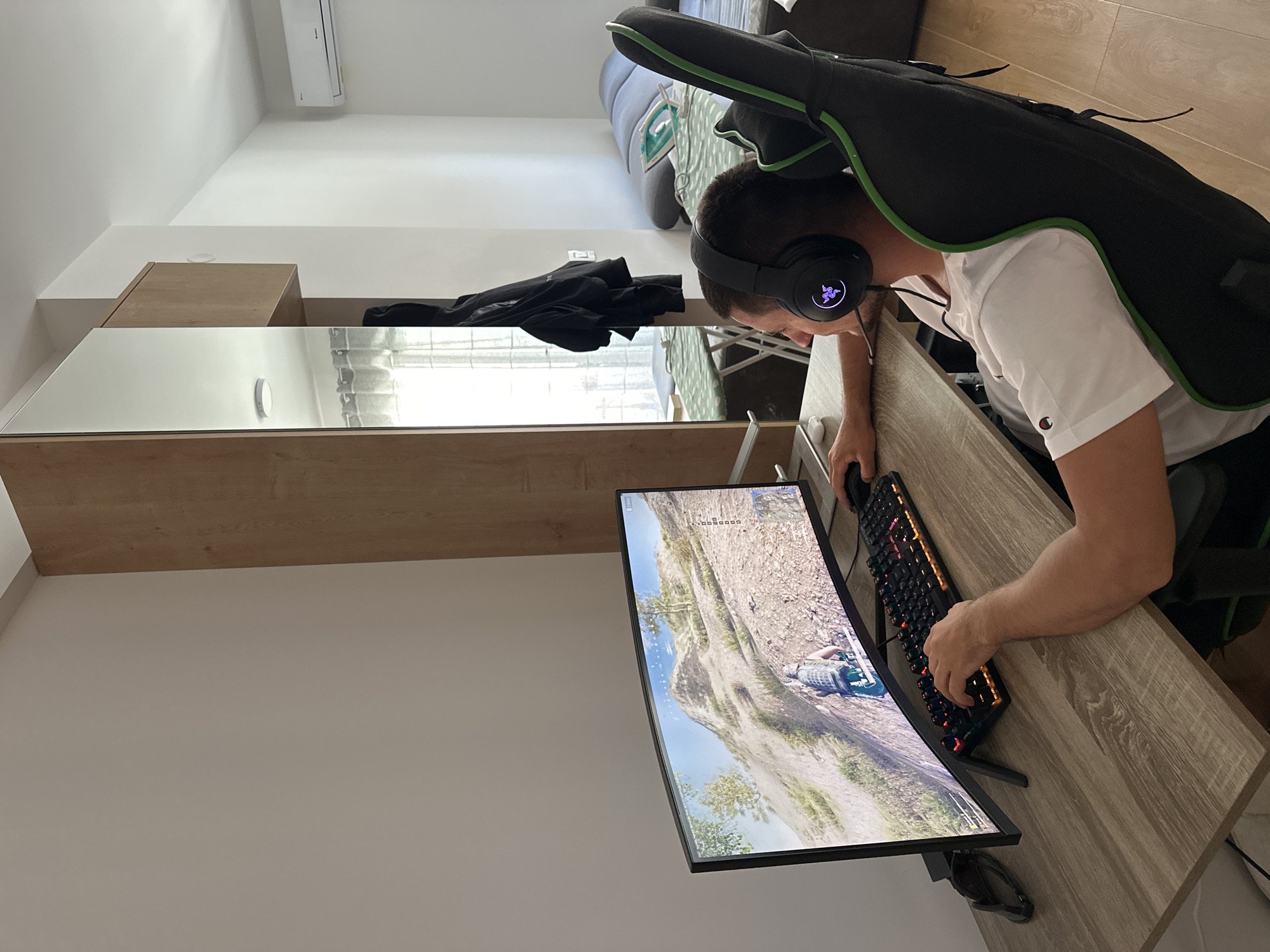 A man is playing a video game on a curved monitor. He is wearing a white t-shirt, headphones, and has a guitar case on his back. He is sitting at a desk with a keyboard and mouse. The game appears to be set in a mountainous or hilly area. The environment includes a light-colored wall, a cabinet with a mirrored surface, and furniture in the background. The location is Donji Pobori, Montenegro.