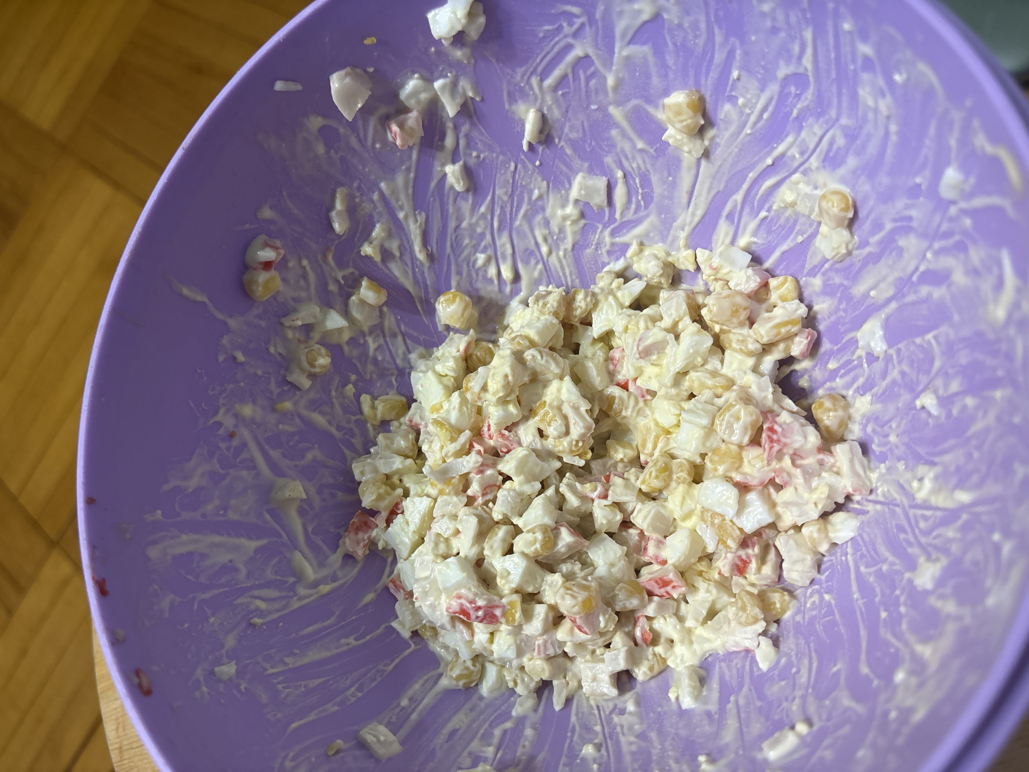 The image shows a close-up, overhead view of a nearly-empty purple bowl containing the remains of a salad. The salad appears to consist of diced imitation crab meat, corn kernels, and possibly hard-boiled eggs, all mixed with a creamy sauce, likely mayonnaise. Traces of the salad mixture are smeared on the inside of the bowl, indicating it has been eaten. The bowl is placed on a wooden surface.
There is no indication of the location being Murino, Russia.