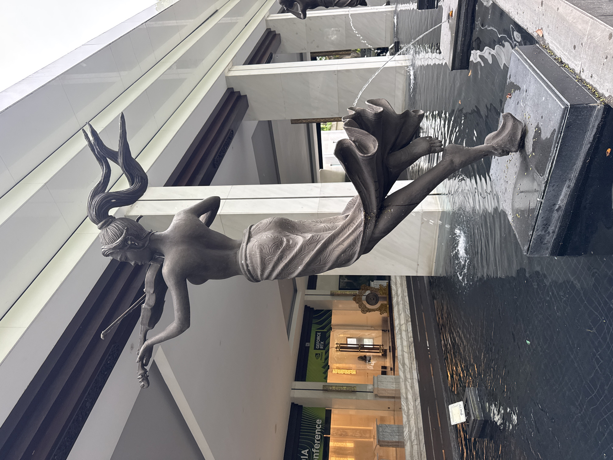 The image captures a statue of a woman playing the violin, positioned above a pool of water. The statue is part of a fountain feature, with water cascading from the top of the statue. The statue is the main subject of the photograph. It is in front of a modern building with geometric architectural elements and a mostly white color. The statue is located at the Renaissance Bali Uluwatu Resort & Spa in Kuta Selatan, Indonesia. In the background are signs for an "NVIDIA Conference" and "GEFORCE RTX". The environment seems to be an entrance or lobby area.