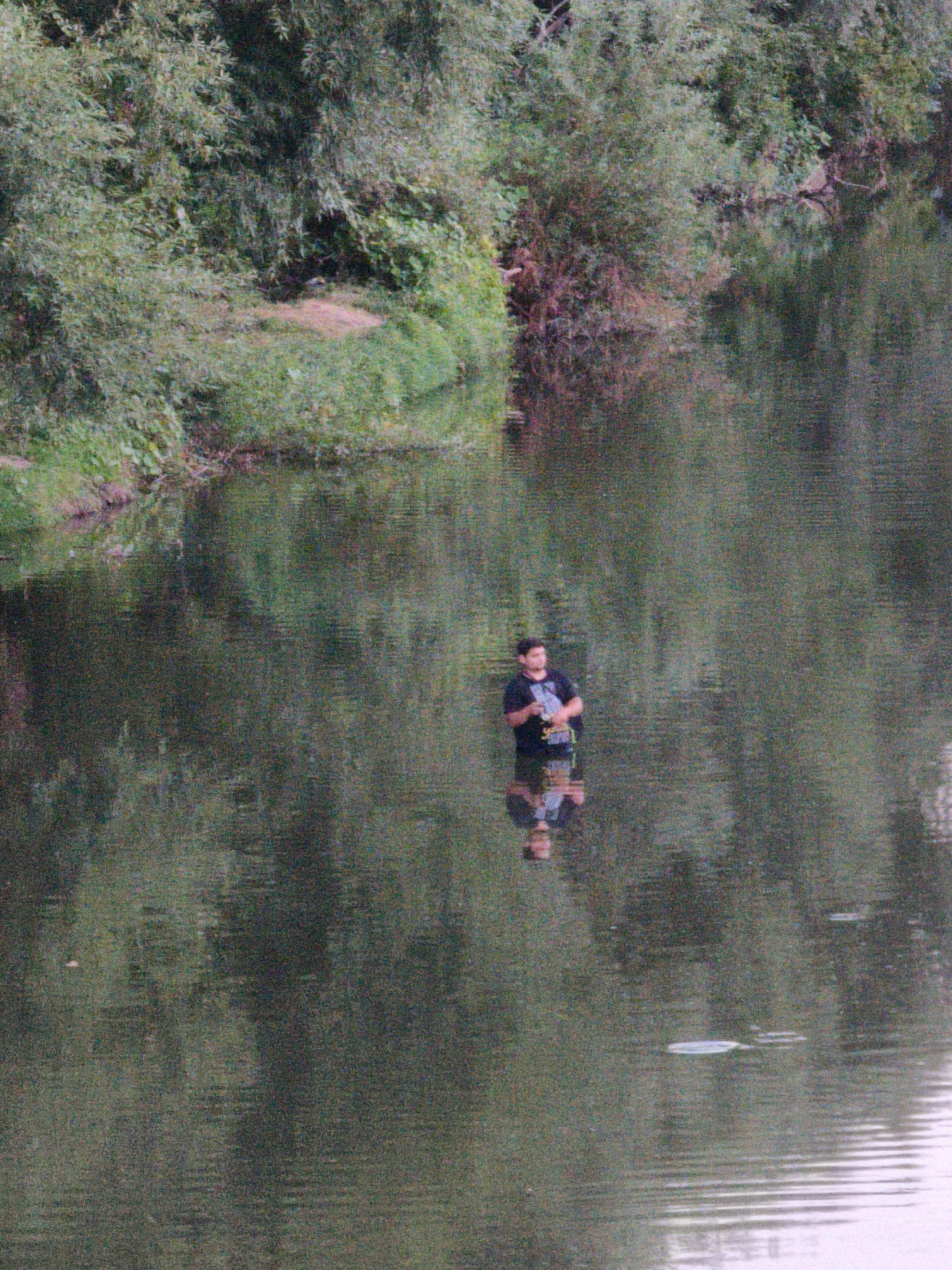A person stands in the shallow water of a river, facing towards the right side of the frame. The person is wearing a dark t-shirt and dark shorts, with their lower legs submerged in the water. Their reflection is visible in the rippling surface. The riverbank on the left is densely covered with green foliage and trees, with a small patch of reddish-brown earth visible on the bank. The water is a dark green color, reflecting the surrounding vegetation. The scene appears to be a natural setting, possibly in Lovech, Bulgaria, given the mention of the city. There are no other people or significant objects in the immediate vicinity. The overall impression is one of a solitary moment in a natural environment.
