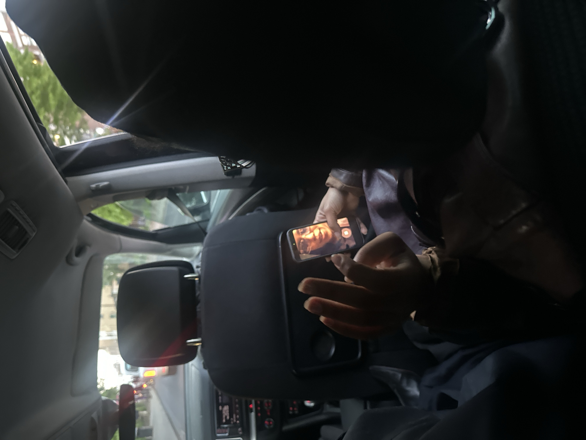 The image depicts the interior of a vehicle, likely a van or a large car, viewed from a rear passenger's perspective looking towards the front. A person, presumably the driver or front passenger, is visible from the chest down, wearing a brown leather-like jacket over a darker garment. They are holding a smartphone in their right hand, the screen illuminated and displaying a man's face, possibly from a video call, a recorded video, or a selfie. A camera shutter icon is also visible on the phone's screen. Their left hand rests on a dark gray headrest or armrest-like structure.
Through the vehicle's windows, an urban outdoor environment is visible, featuring green trees and buildings, consistent with a London setting. A sunroof or open roof panel is visible at the top, offering a glimpse of the sky and foliage. A large side mirror on the left reflects traffic or streetlights, indicating other vehicles on the road. The dashboard and some controls of the vehicle are faintly discernible below the armrest. A small device, possibly a dash cam, is attached near the top center of the windshield area.
The scene suggests the person is engaged with their phone while in transit. The interior appears somewhat dark, contrasted by the brighter ambient light from outside and the glow of the smartphone screen. The time of day appears to be daylight, possibly late afternoon or early evening. No distinct text is visible on any objects or screens.