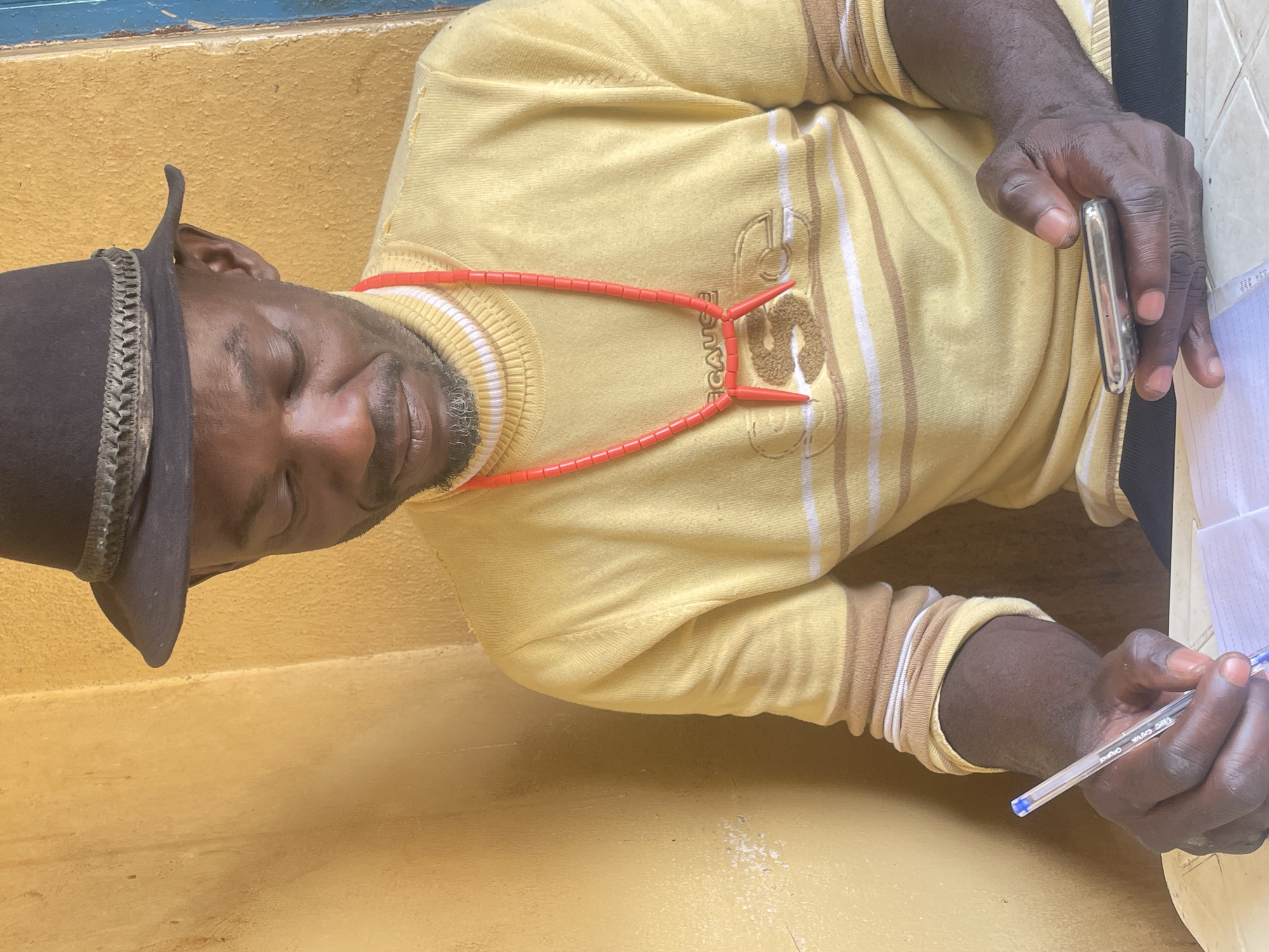 A man is sitting on a painted yellow step, wearing a brown hat, a yellow sweater with stripes and text, and a red necklace. He is holding a pen and a metal object and appears to be writing on a piece of paper. The scene is presumably located in Jos, Nigeria.