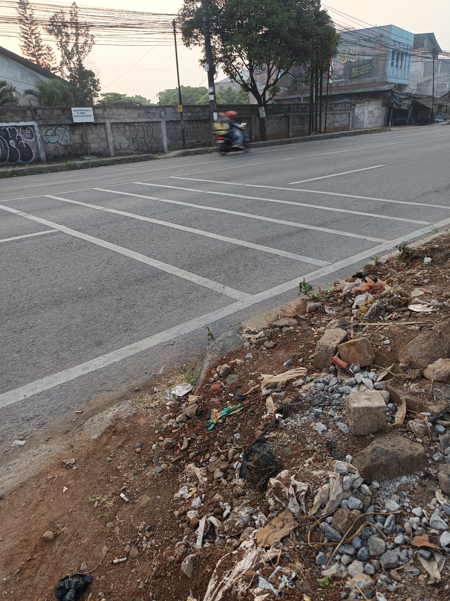 The image depicts a street scene in Talaga Sari, Indonesia, focusing on the roadside. A paved road with white lane markings runs horizontally across the frame. On the left side of the road, a concrete wall is visible, adorned with graffiti. Beyond the wall, trees and a building are partially obscured. A person on a motorcycle is in motion on the road, appearing blurred due to movement. The right side of the road is a dirt embankment littered with various types of debris, including plastic bags, rocks, and what appear to be discarded packaging and cigarette butts. The ground is uneven and covered in loose soil and stones. Power lines are strung across the upper portion of the image, and the sky appears overcast or hazy. The overall impression is one of urban decay and neglect in this specific area.