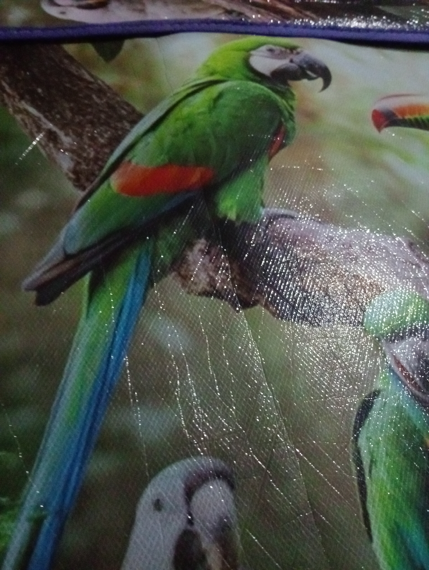 The image shows a close-up of fabric with a printed design of several birds. The dominant bird is a green macaw with a red patch on its wing and blue tail feathers, perched on a branch. Another green macaw is partially visible at the bottom right. A grey-and-white bird is partially visible at the bottom center of the image. In the background, to the upper right of the main macaw, part of a toucan is visible. The fabric has a shiny, textured surface that obscures some of the details. The exact location in Guhauleu, Indonesia, cannot be determined from this image.
