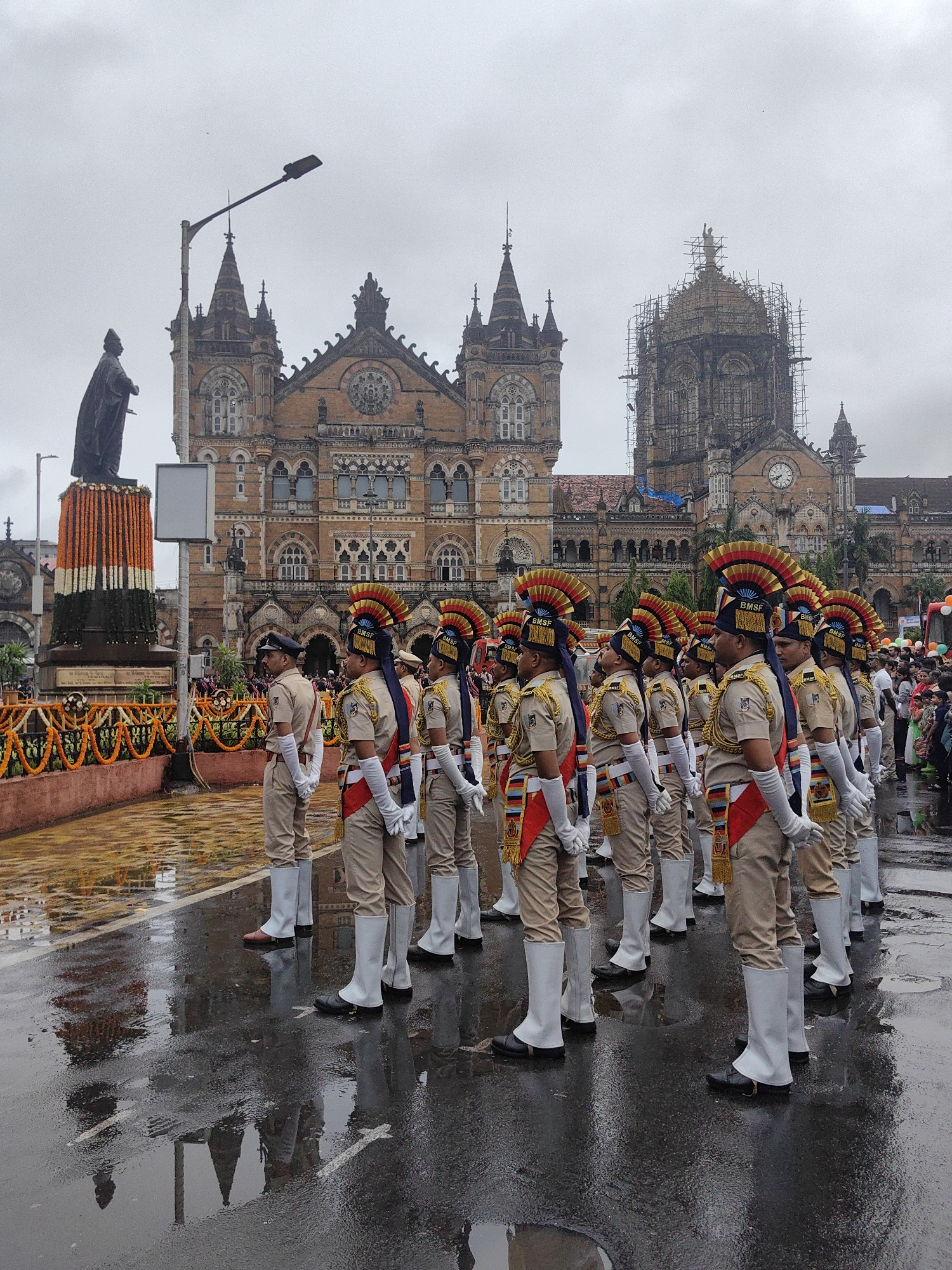 The image depicts a solemn ceremonial procession in front of the Chhatrapati Shivaji Maharaj Terminus (CSMT) in Mumbai, India. A line of uniformed individuals, dressed in ceremonial attire including tall white boots and elaborate headdresses, stand at attention on a wet, reflective surface that appears to be a paved street or plaza. Behind them, CSMT, a UNESCO World Heritage site known for its Gothic Revival architectural style, serves as a grand backdrop. The building's intricate facade is visible, with one of its domes partially covered in scaffolding, suggesting ongoing restoration or maintenance. The sky is overcast, contributing to a subdued atmosphere. In the foreground, to the left, a statue stands on a pedestal adorned with orange, white, and green garlands, possibly representing the Indian flag. The wet ground reflects the surroundings, creating a mirror-like effect that adds depth to the scene.