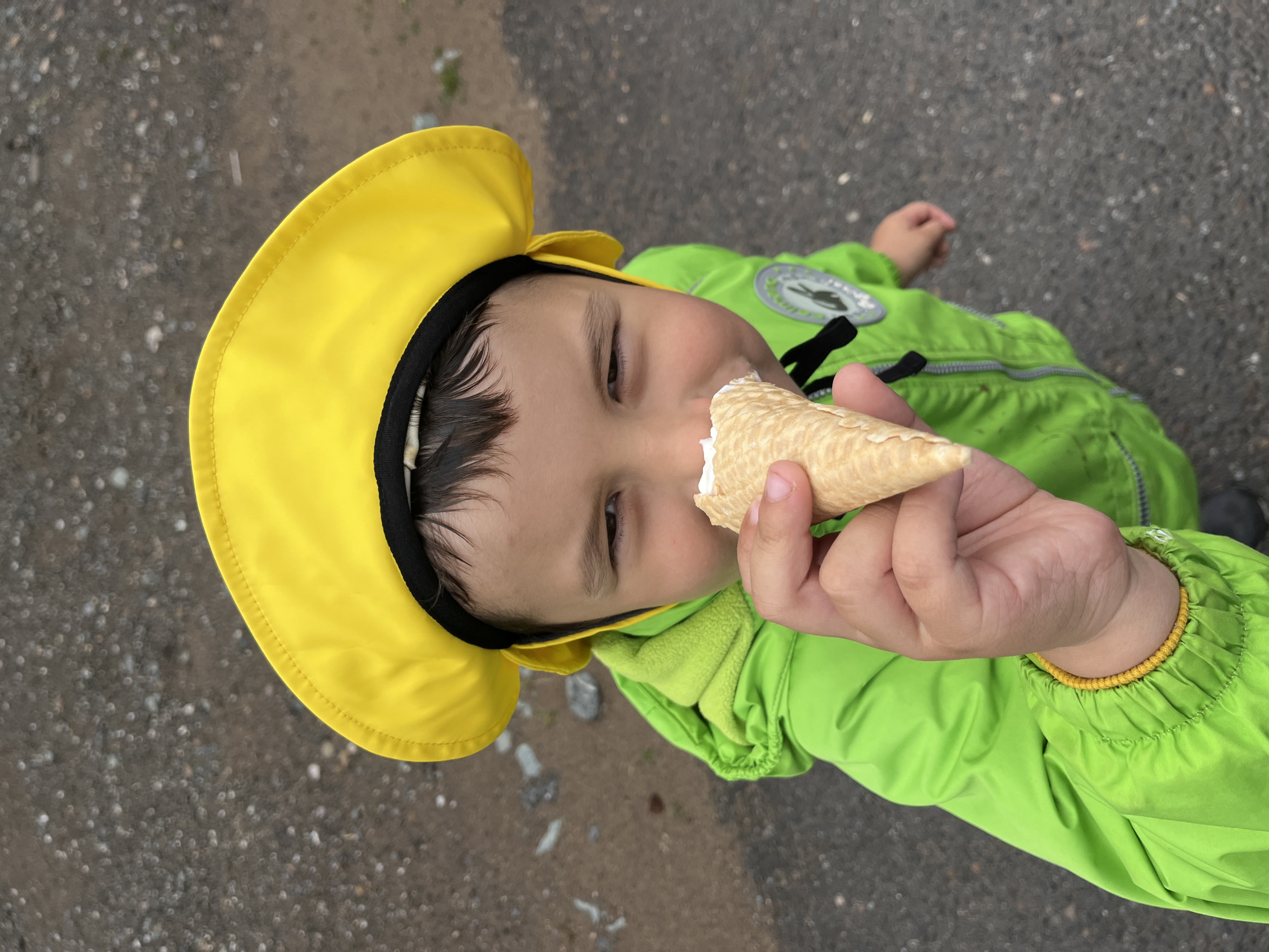 The image is a close-up, high-angle shot of a young boy wearing a bright yellow sun hat and a neon green jacket. He is holding an ice cream cone to his mouth, with a small amount of white ice cream visible on the cone. The boy's face is partially obscured by the ice cream cone, but his eyes are visible, looking towards the camera. The background consists of a textured, gray surface, likely asphalt or gravel, with some scattered small rocks and pebbles. The lighting suggests an overcast or outdoor setting. The boy's clothing has a black trim on the hat and a logo on the jacket. A yellow elastic band is visible on the cuff of his jacket. The overall scene appears to be a candid moment captured outdoors, possibly in a park or on a street in Chaikovsky, Russia.