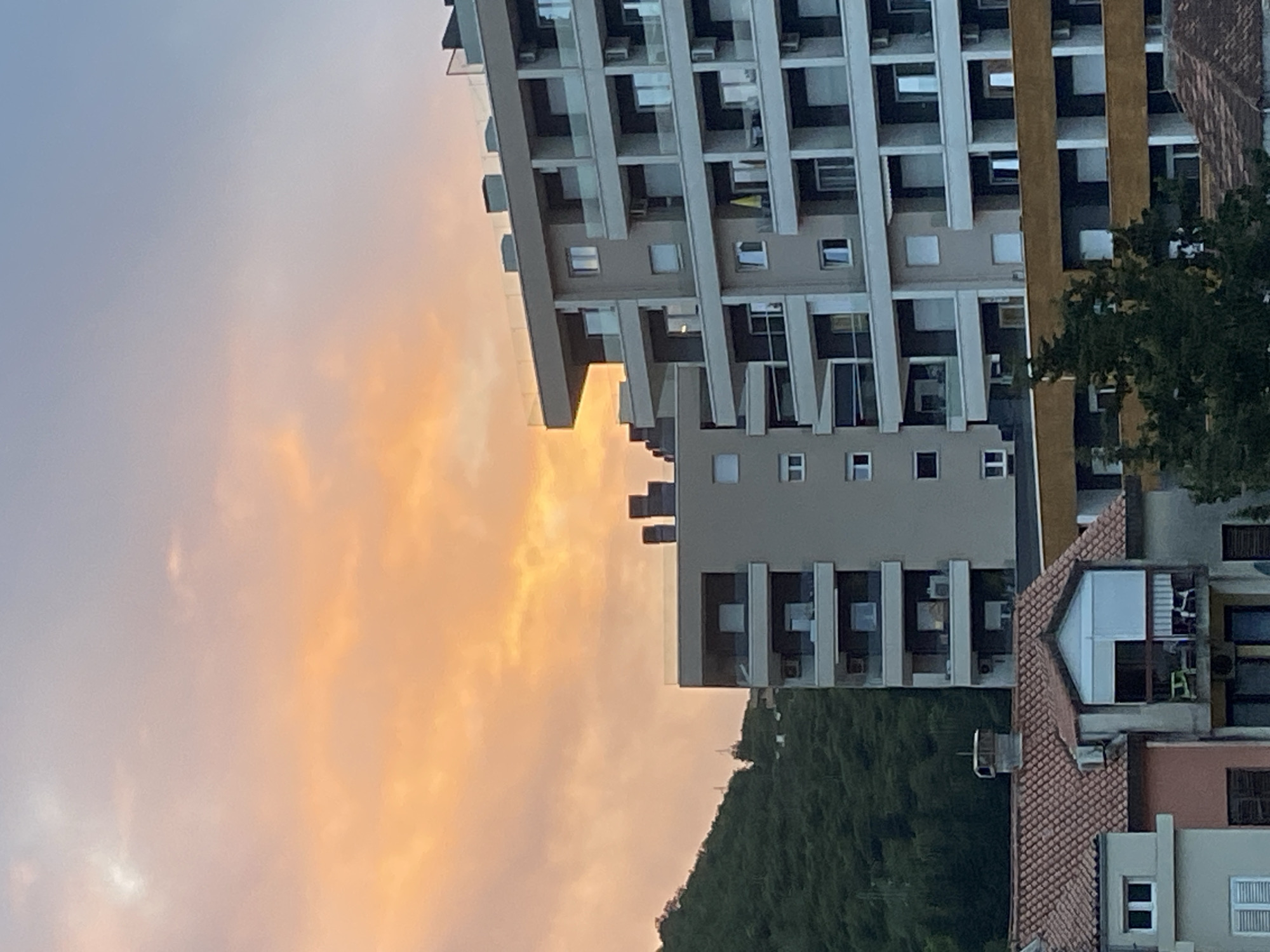 The image shows a view of buildings in Budva, Montenegro, at sunset. A tall, modern building with multiple balconies dominates the upper right portion of the frame. Adjacent to it are other buildings with red-tiled roofs. In the lower center, there is a forested hillside. The sky is filled with clouds, tinted with orange and pink hues from the setting sun.