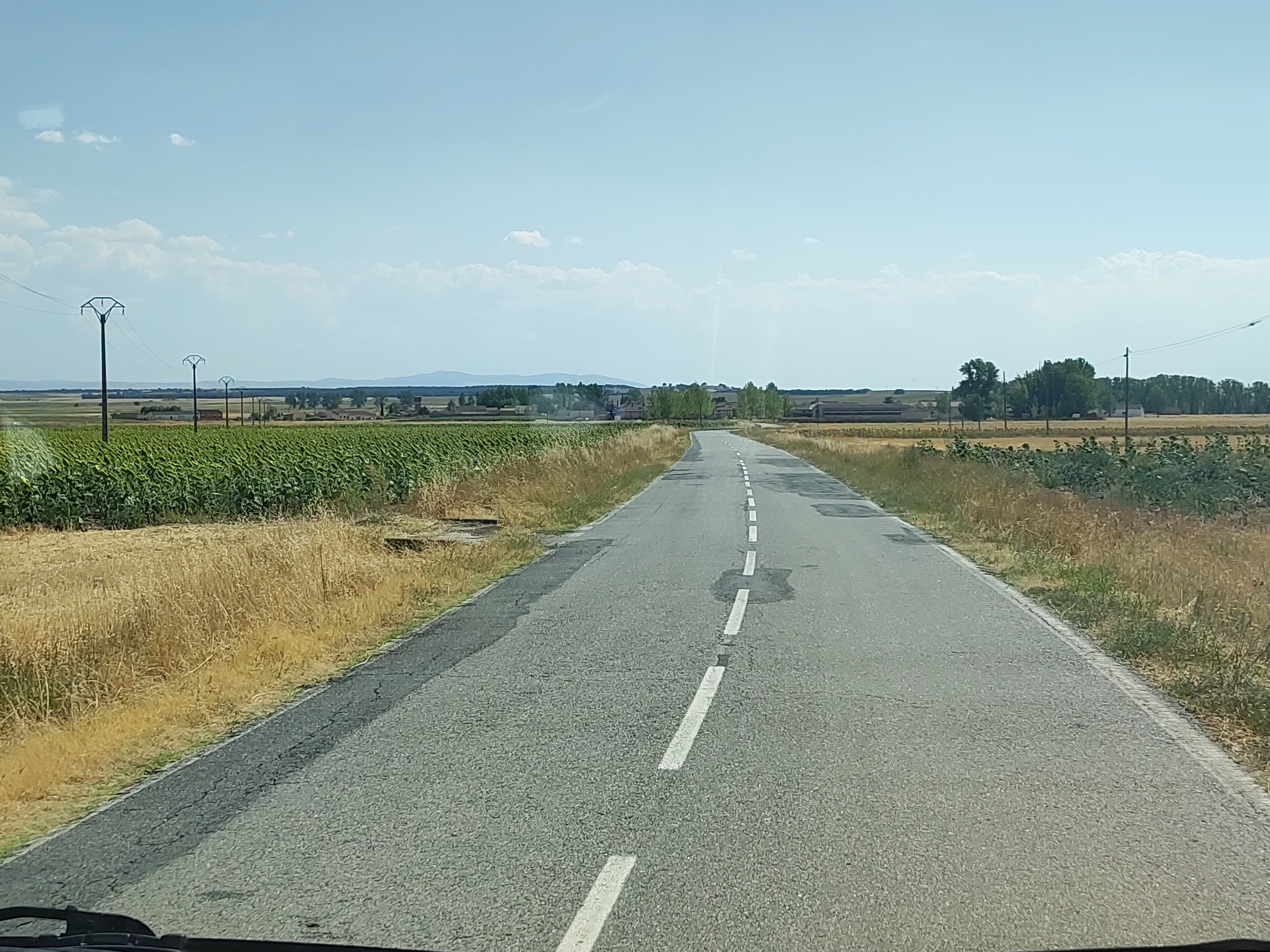 The image shows a view from inside a vehicle looking forward along a paved road in Santa María la Real de Nieva, Spain. The road has a broken white line down the center and appears to be asphalt. On either side of the road, there are fields. The left side shows a field of green crops, possibly sunflowers, and dry, yellow grass. The right side has fields of dry, yellow grass and some green vegetation. In the distance, there are trees and some buildings, suggesting a rural or semi-rural setting. Power lines and poles are visible in the distance on both sides of the road. The sky is clear with a few scattered white clouds. The overall scene depicts a typical rural landscape on a sunny day.