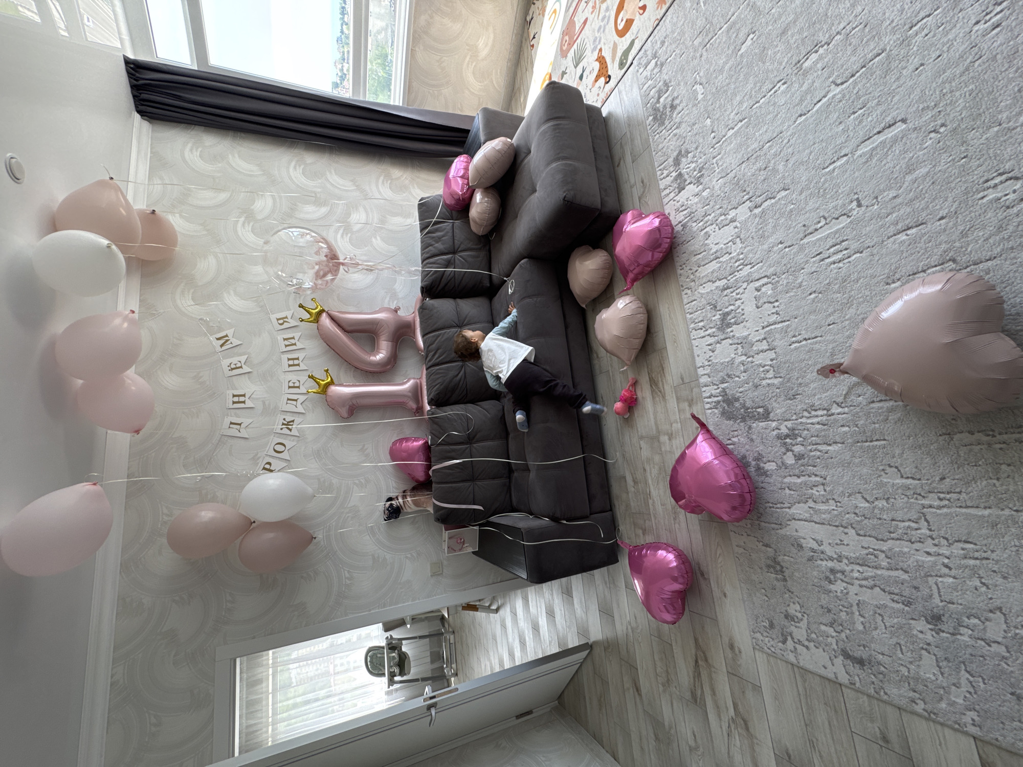 A high-angle, indoor shot captures a room decorated for a birthday celebration, likely in Sochi, Russia, given the Cyrillic script on the "Днем рождения" banner. A grey modular sofa is the central element, adorned with pink and white heart-shaped balloons and number-shaped foil balloons suggesting a 14th birthday. A young child is lying on the sofa. The room has patterned wallpaper, a grey carpet, and light wood flooring. A doorway is visible in the lower left corner, leading to a glimpse of what seems to be a baby seat. The room is well-lit, likely by natural light from a window partially covered by a dark curtain.