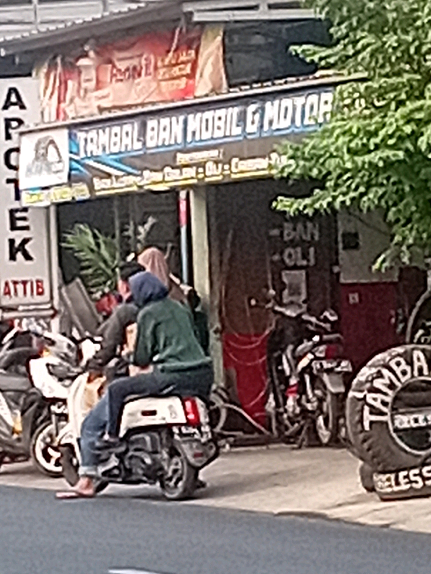 In Besito, Indonesia, a scene unfolds outside a shop with a prominent sign reading "TAMBAL BAN MOBIL & MOTOR" (Tire Repair for Cars & Motorcycles). The shop appears to be a service center, with several motorcycles parked outside. Two individuals are seated on a white scooter in the foreground. The person in front, wearing a green hooded jacket and jeans, is operating the scooter. The person behind, wearing a hijab and a lighter-colored garment, is also seated on the scooter. A large tire with the inscription "TAMBAL" is visible to the right of the scene, along with other motorcycles and some foliage. The shop's facade displays additional signage and what appears to be internal shelving. The overall environment suggests a commercial street scene in a town or city.