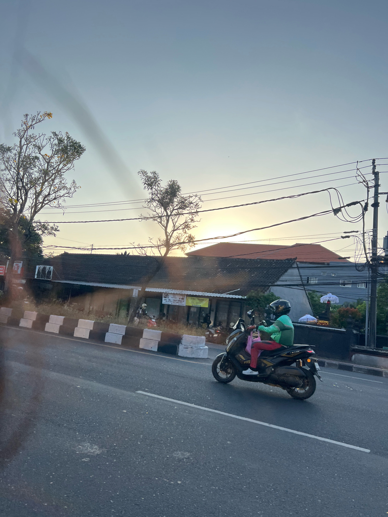 The image captures a street scene at dusk in Denpasar, Indonesia. A person is riding a black scooter on the right side of the road, wearing a green jacket, red pants, and a helmet. The scooter has a license plate that is not legible. In the background, there are buildings with tiled roofs, power lines, and trees. The sun is setting, casting a warm glow over the scene. The road surface is asphalt with a white dividing line. Along the side of the road, there are concrete barriers and some vegetation. The overall impression is a typical urban street during the evening.