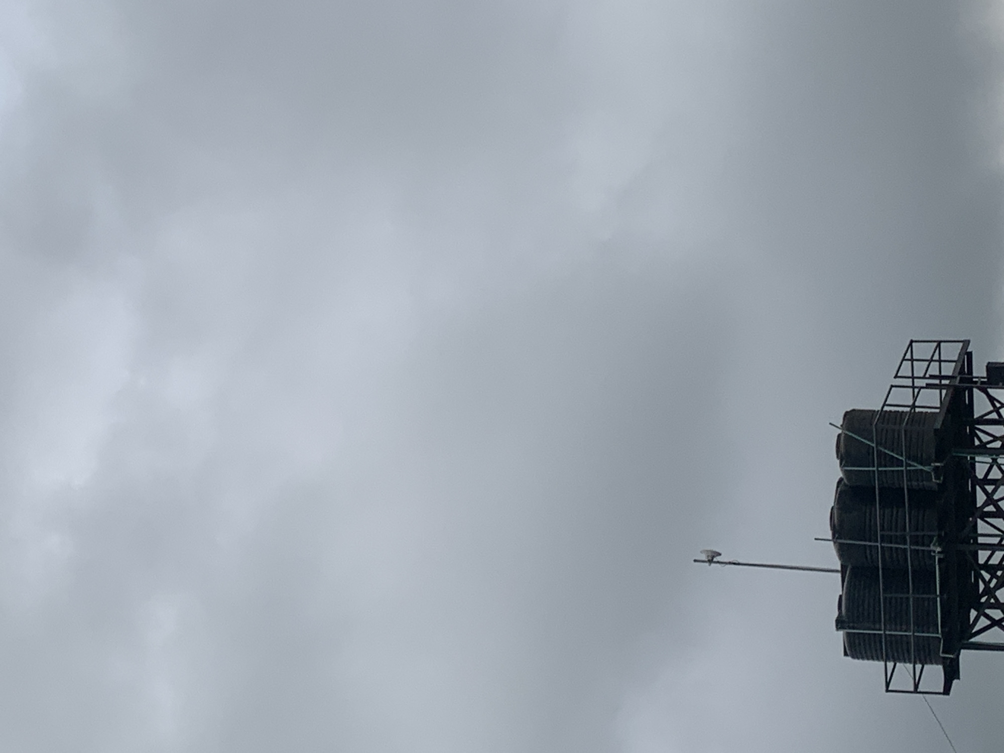 In the city of Unknown, Unknown, under a cloudy sky, a close-up shot focuses on a water tank tower. The tower's dark, cylindrical tanks are stacked on a metallic structure with a railing. A thin antenna stands tall behind the tanks, topped with a white, directional device. The overcast sky dominates the background, creating a muted and diffused light across the scene.