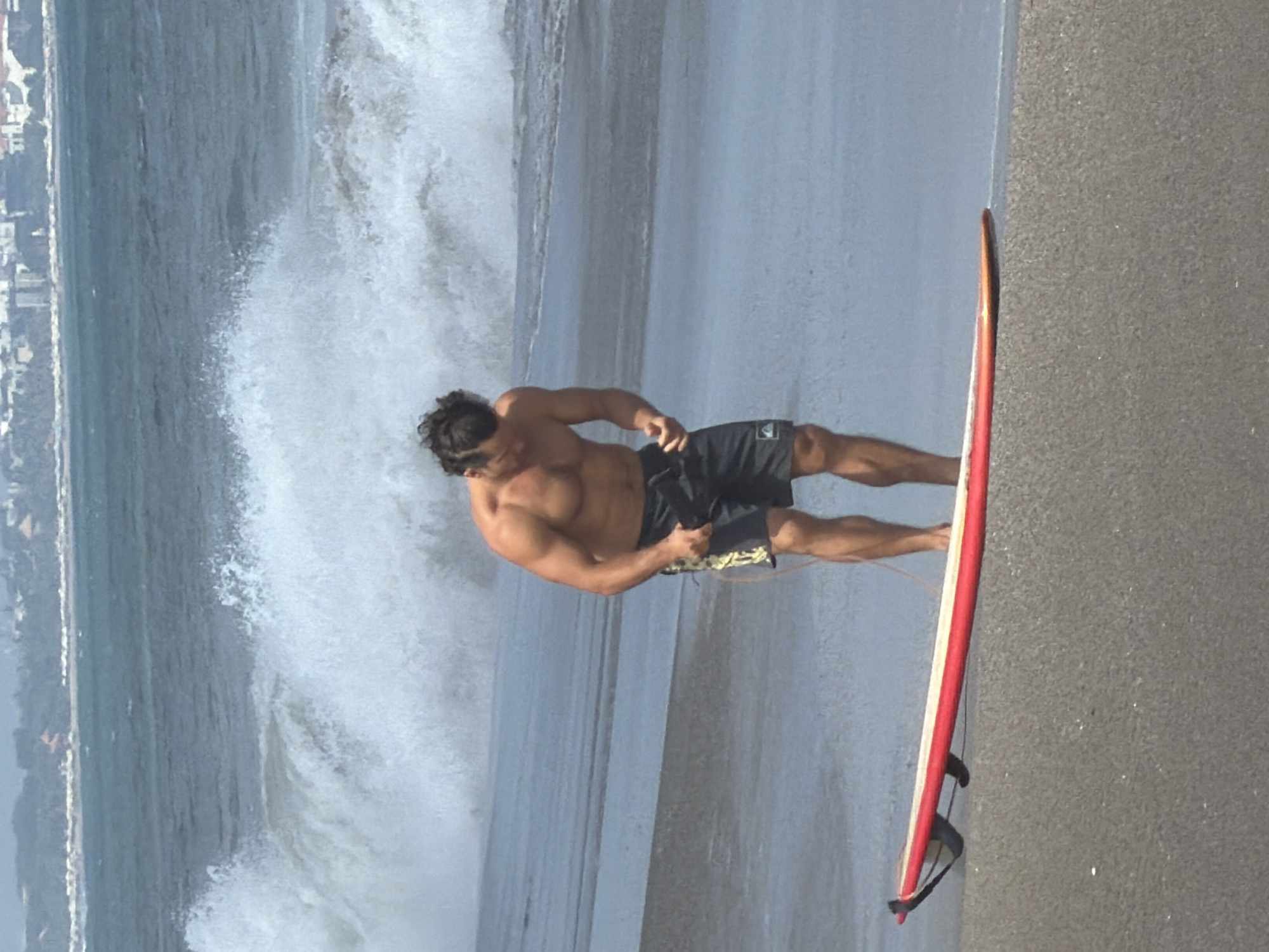 In North Kuta, Indonesia, a shirtless man with dark curly hair stands on a sandy beach next to a red surfboard. He is wearing black swim shorts with a yellow pattern. The man is looking down at his hands, which are clasped together. Behind him, a large wave is crashing in the ocean, creating white foam and spray. In the distance, a cityscape with buildings is visible along the coastline. The overall scene depicts a moment of anticipation or reflection on the beach.