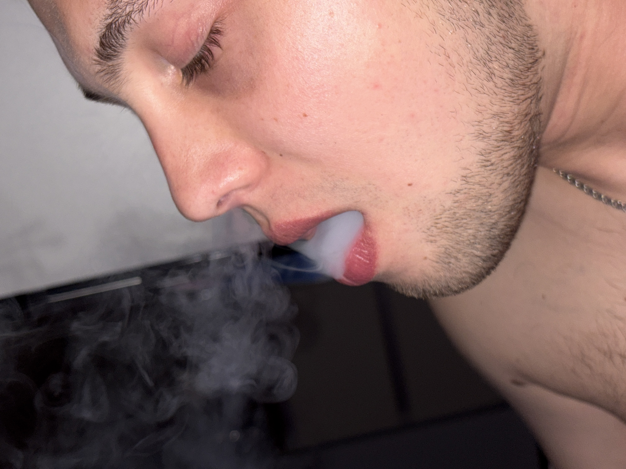 A close-up image shows a person exhaling a cloud of smoke. The person has light skin, dark hair, and a short beard. The camera angle is from the side, focusing on the person's face, particularly their nose, mouth, and jawline. A silver chain is partially visible around their neck. The background is blurred.