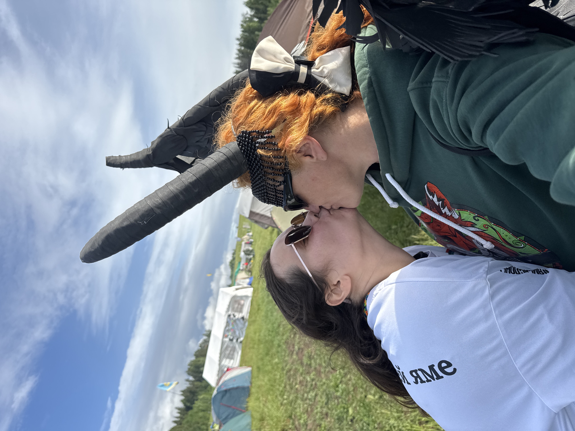 A sideways-oriented outdoor photograph captures two individuals kissing under a bright, cloudy daytime sky. The scene appears to be at a festival or camping event in Большая Кивара, Russia, with a grassy field and numerous tents in the background.
The person on top has vibrant reddish-orange curly hair, adorned with a black and white bow hair clip and a black beaded facial accessory across their forehead. They wear a dark green hooded sweatshirt featuring a colorful graphic design, possibly a creature, on the chest. Elaborate black, possibly faux-leather, horns extend upwards from behind their head, complemented by black feathers visible on their right shoulder.
The person on the bottom has long dark brown hair and wears sunglasses perched on their nose. They are dressed in a white t-shirt, which displays black text. Partially visible text reads "Й ЯМЕ," and on the sleeve, the reversed hashtag "#ОНЛАЙН_В_ЯМЕ" is discernible. A subtle colorful, possibly rainbow-colored, strap is visible near their neck.
In the background, various white and blue tents are scattered across the green landscape, with distant trees marking the horizon. A yellow and blue flag is also visible against the cloudy sky. The image conveys a moment of affectionate intimacy amidst an outdoor gathering.