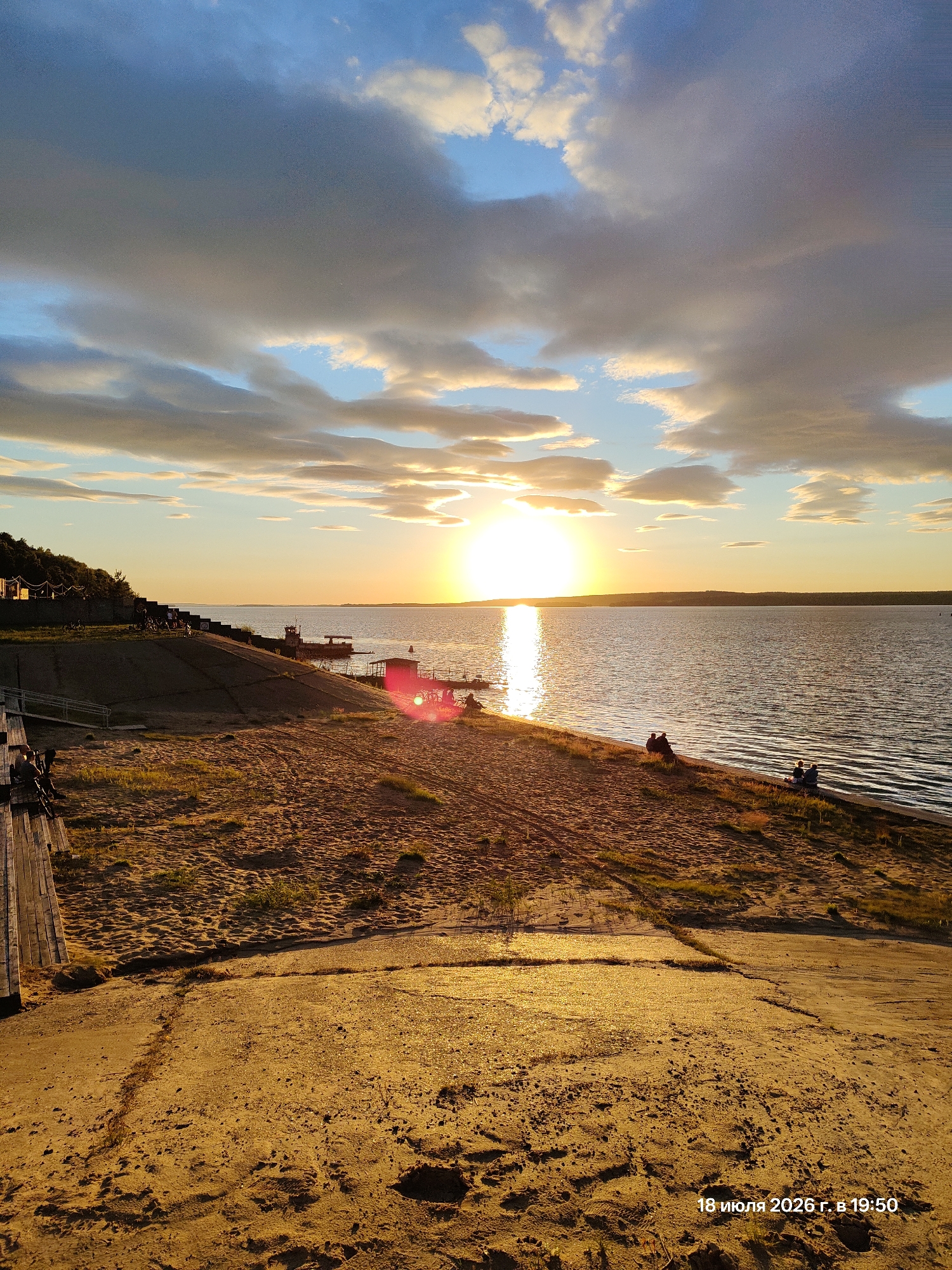 This is a photograph taken at sunset on July 18, 2026, at 7:50 PM. The image depicts a sandy beach area adjacent to a large body of water, likely a river or lake. The sun is setting on the horizon, casting a warm glow across the scene and creating a reflective path on the water. The sky is a mix of blue with scattered clouds, some of which are illuminated by the setting sun.
Along the shoreline, there are several small structures and a pier. A few people are visible, either sitting on the sand, walking, or interacting near the water's edge. The foreground is dominated by sand and some dry grass. A wooden structure, possibly a boardwalk or bench, is visible on the left side of the image. The background features a tree-covered hill with some buildings or tents at the top, suggesting it might be a recreational or residential area. The overall atmosphere is peaceful and scenic, characteristic of a summer evening.