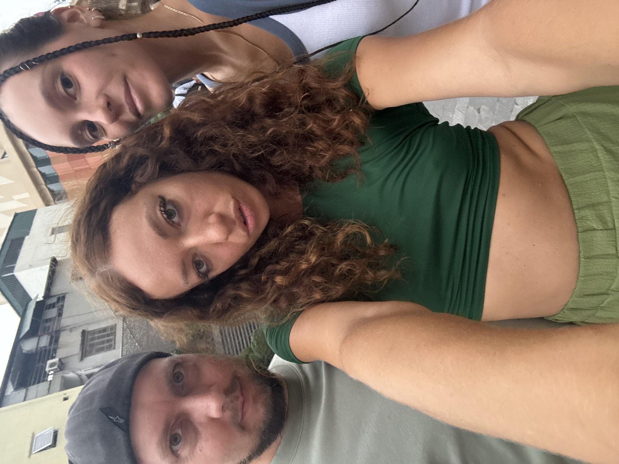 A selfie-style photograph shows three individuals in an urban environment, identified as Hà Nội, Vietnam by the background architecture and signage. The individuals are close to the camera, creating a crowded composition. A woman with braids and multiple ear piercings is positioned towards the top of the frame, wearing a white tank top with blue accents. A woman with curly brown hair and a green cropped top and green bottoms is centrally located, directly facing the camera. A man with a beard and a gray cap is at the bottom, smiling slightly.
The background shows buildings with architectural details characteristic of the region, including window styles and construction materials. There is also signage present, presumably in Vietnamese. The image suggests a casual, tourist-style photograph taken during the daytime.