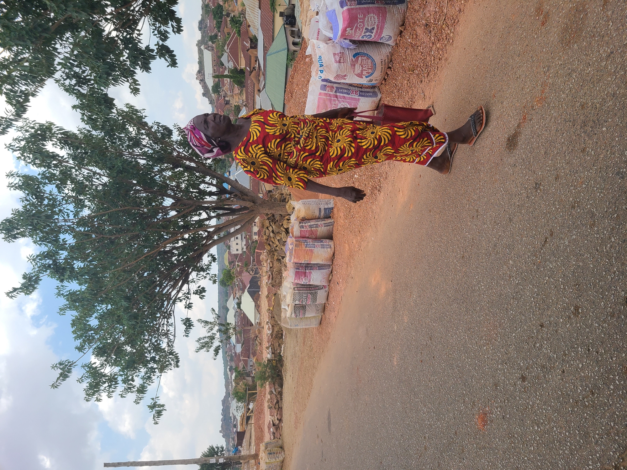 The image shows an elderly woman walking on the side of a paved road in Jos, Nigeria. She is wearing a colorful patterned dress and sandals, with a head wrap and carrying a red bag. To her right, bags of cement are stacked on a dirt area beside the road. In the background, a residential area with various buildings is visible, and a tree stands to the left of the woman. The sky is partially visible behind the tree.