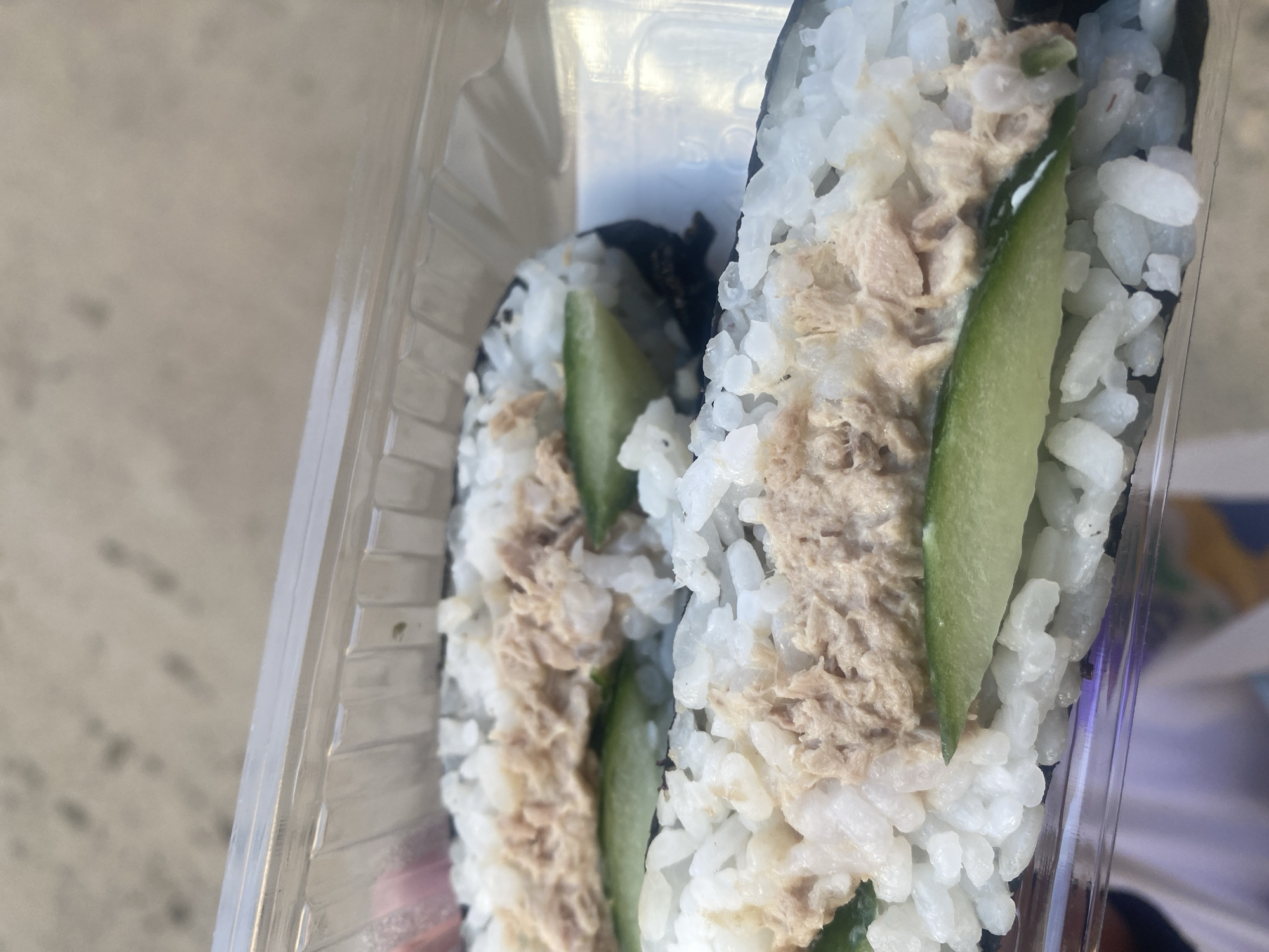 The image shows a clear plastic container holding two sushi rolls. The rolls are made with white rice, seaweed, tuna, and a slice of cucumber. The location appears to be indoors, with a blurry background.