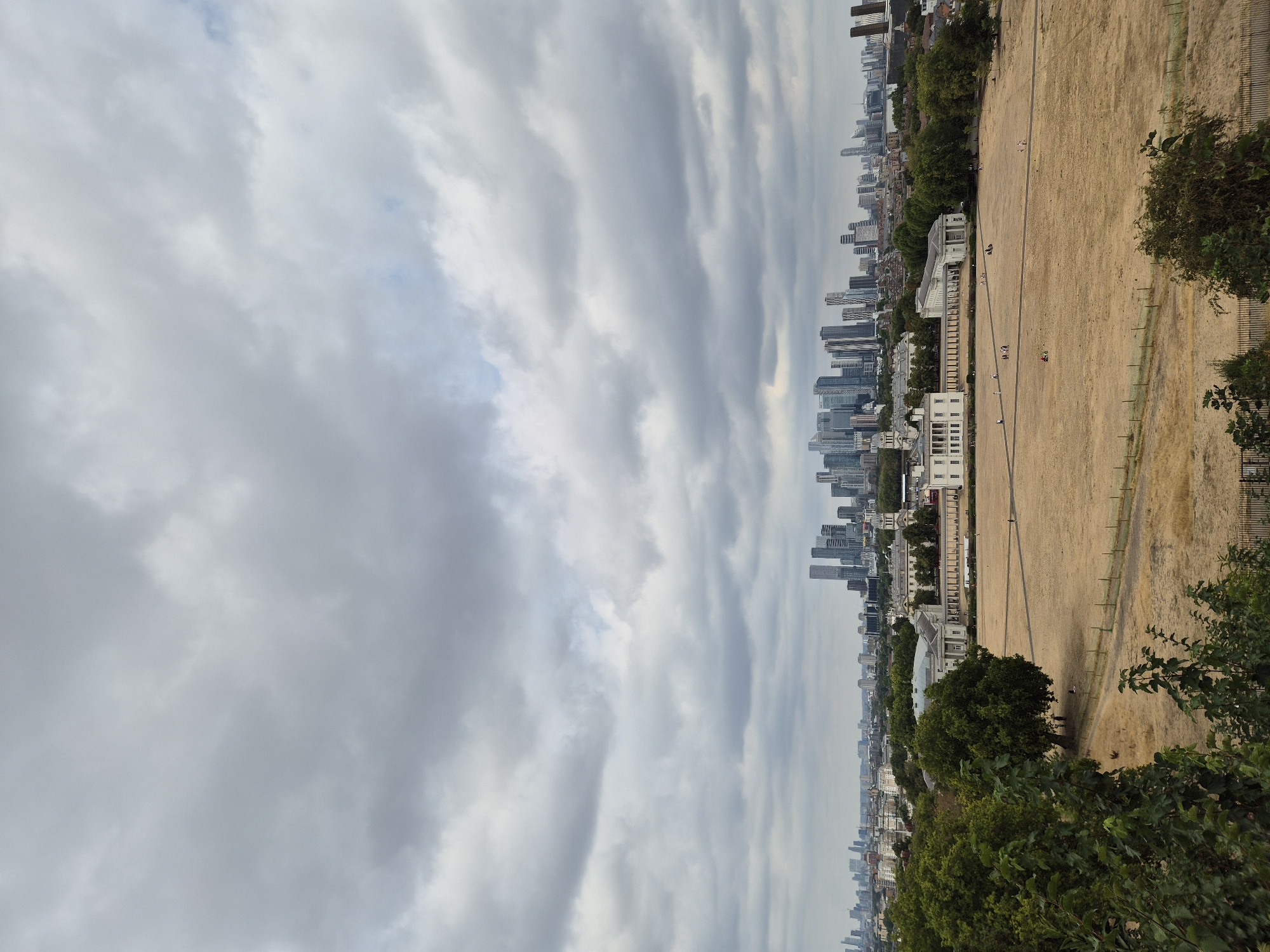 The image is a wide shot of a park in London, United Kingdom, on a cloudy day. The foreground shows lush green trees and foliage. In the midground, there is a large, dry, grassy field with a few scattered people walking and sitting. Several white colonial-style buildings with columns and arched windows are situated at the edge of the field. In the background, a dense cityscape with numerous modern skyscrapers dominates the horizon, suggesting a central business district. The sky is overcast with heavy gray clouds.