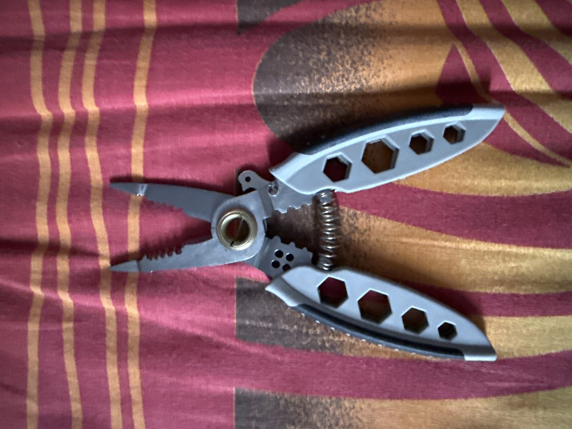 The image shows a pair of gray and silver multi-pliers lying open on a red and yellow striped fabric. The pliers have serrated jaws and a coiled spring in the center. The fabric has a pattern of horizontal stripes, with some darker, blurred shapes visible beneath them. The scene is well-lit and the object is in focus. There are no people or animals visible in the image. The location of Морозовка, Russia is not depicted in the image.