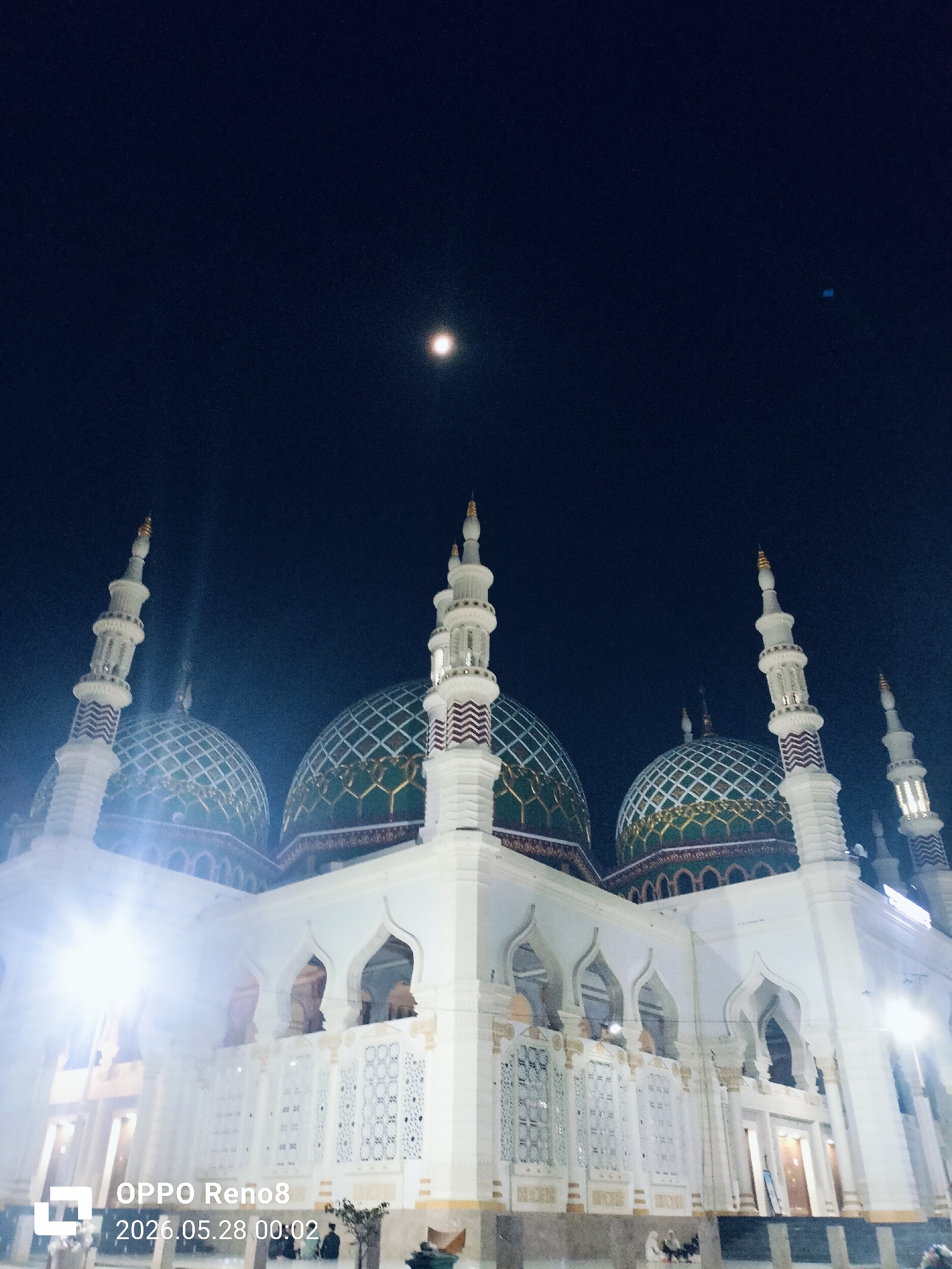 The image captures the Baiturrahman Grand Mosque in Kotapantonlabu, Indonesia, at night. The mosque's architecture features white walls, arched openings, and domes with a green patterned design. Several minarets with white structures and striped details are visible. The sky is dark, with a visible moon. A light source illuminates the mosque from the bottom left. There are people sitting and standing at the base of the mosque. A "OPPO Reno8" watermark is present in the lower-left corner, along with the date "2026.05.28 00:02".