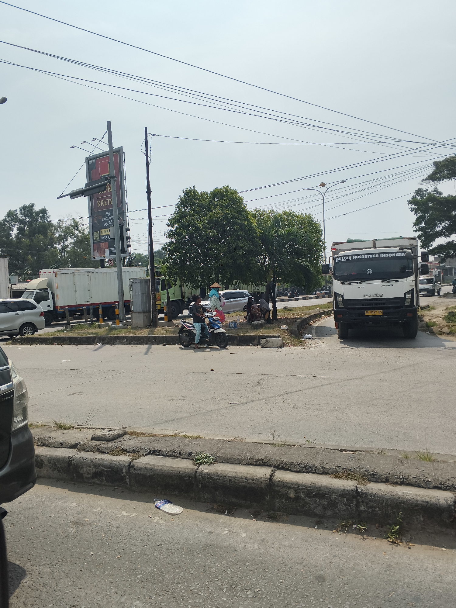 The image depicts a street scene in Bandar Lampung, Indonesia. In the foreground, the edge of a dark-colored car is visible on the left. The road surface is asphalt, with a concrete curb and median strip. A single white flip-flop lies on the road near the curb. Further into the scene, a white truck with a closed cargo box is parked on the right side of the road. The truck has "ISUZU" written on its front and Indonesian license plates. A white truck with a similar cargo box is parked to the left of it. In the background, several trees are visible, including a cluster of tall, leafy trees. A large billboard with red and white signage for "KRETEK" is present near the center. A person wearing a conical hat and a blue headscarf is walking a motorcycle with a passenger. Other individuals are seated near the trees. Electrical wires crisscross the sky, and a streetlamp is visible. The overall impression is of a busy urban environment with commercial activity and pedestrian and vehicular traffic.