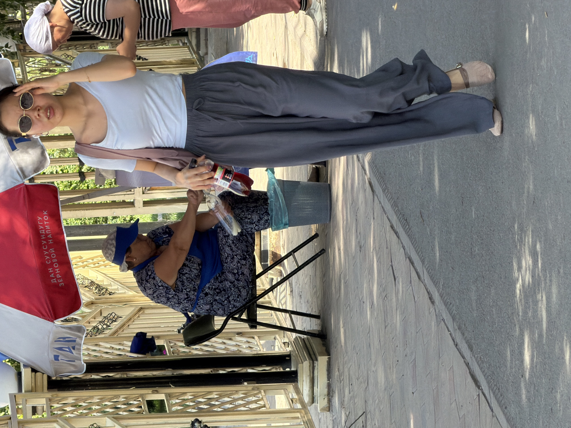 A medium shot captures an outdoor scene on a paved walkway adjacent to a street, with dappled sunlight illuminating the ground. In the foreground, a young woman walks away from the camera, her left hand holding a bottle of red liquid. She wears a light blue crop top, loose gray pants tied at the waist with a drawstring, and flat, light-colored shoes. Her right hand is raised to adjust her oval-shaped sunglasses. To the left of the young woman, a seated woman wears a dark patterned dress, a blue apron, and a blue visor. She is leaning forward, her hands engaged with a transparent plastic bottle. Behind the seated woman is a collapsible chair and a fence. Further back on the right, another woman with a white headscarf and a striped shirt and pink pants stands near a light-colored railing. The background features an umbrella canopy with red and white sections. The environment appears to be a marketplace or street vendor area.