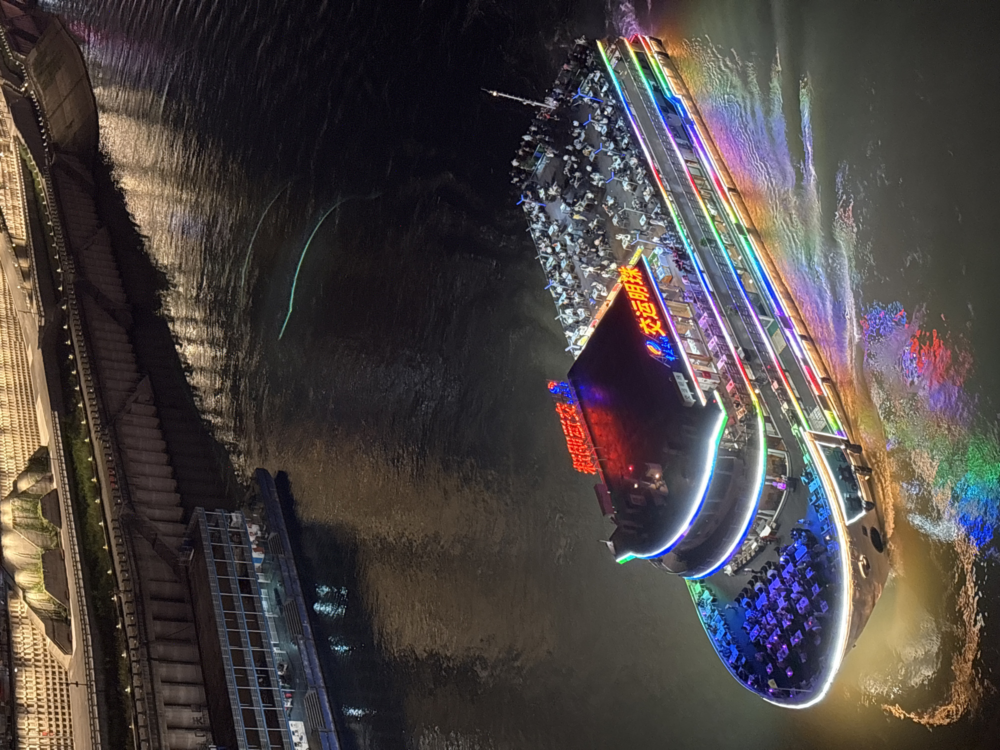 A nighttime view of Nan'an District, China, displays a brightly lit river cruise ship on the Jialing River. The multi-deck ship is adorned with colorful neon lights that reflect in the dark water. People are visible on the ship's open decks, seated at tables, and possibly enjoying dinner or drinks. A concrete embankment with steps and a building are visible on the left side of the image, along the riverbank.