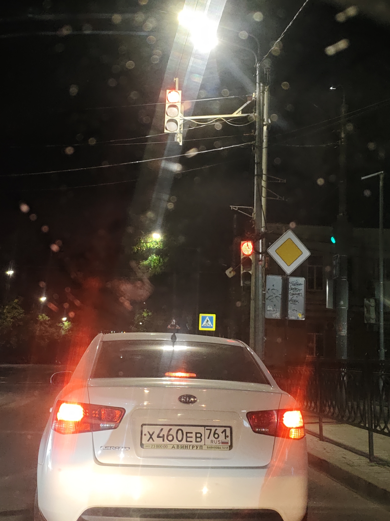 A white Kia Cerato sedan, with license plate X460EB 761, is stopped at a traffic light at night in Rostov-on-Don, Russia. The traffic light is red, and the car's brake lights are illuminated. The scene is viewed from behind the car, looking towards the intersection. The environment appears to be a city street with streetlights and trees visible in the background. There are other cars and traffic signals in the distance. The image quality is slightly degraded by what appear to be water droplets or smudges on the lens, creating circular light artifacts.