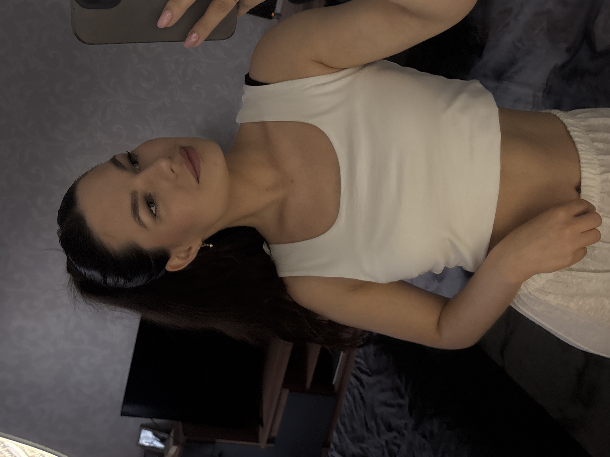 A young woman with her dark hair in a high ponytail takes a selfie in a dimly lit room. She is wearing a white cropped tank top and light-colored pajama bottoms. She is looking at her phone, which she holds in her right hand. A large television is visible in the background, as well as a dresser with a small framed picture on it. The room appears to be a bedroom, with a bed covered in dark bedding visible on the left side of the frame. The wallpaper has a subtle damask pattern. This is a candid shot taken inside a private residence, likely a bedroom, in Chaikovsky, Russia.