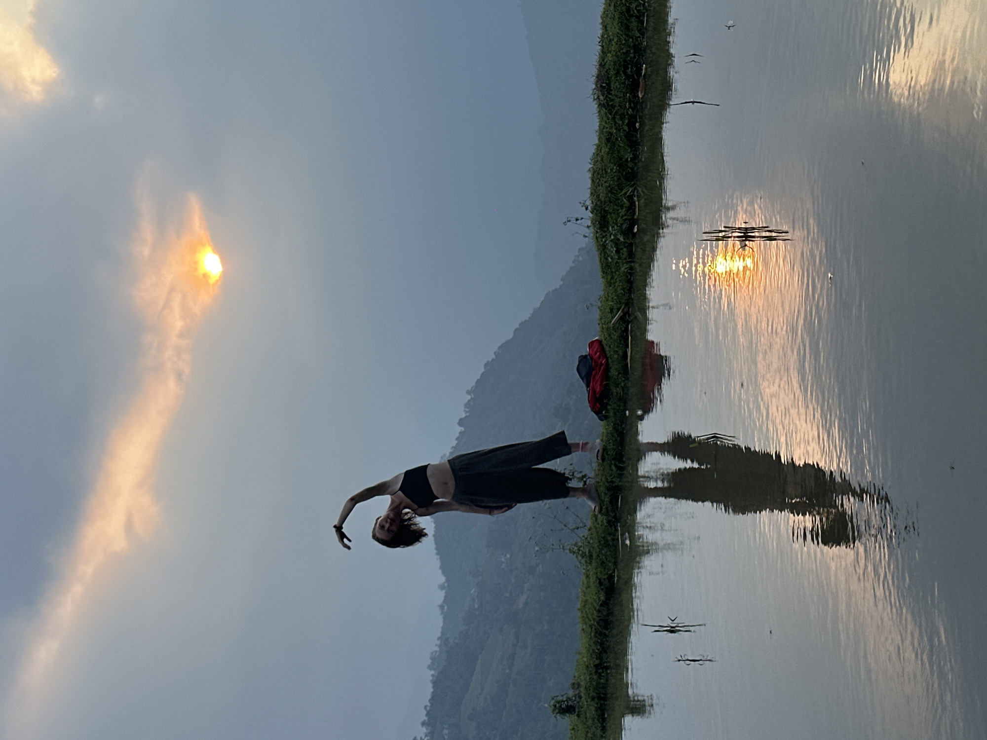 The image captures an ethereal outdoor scene at either sunrise or sunset in a mountainous region, likely Nậm Dịch Commune, Vietnam. The central subject is a woman, standing gracefully on a narrow strip of land, which appears to be a dike bordering a calm body of water, possibly a rice paddy.
The woman has fair skin and dark hair, and is dressed in a dark sports bra and dark wide-legged trousers. She is depicted mid-pose, arching her back and extending her arms upward and outward in a fluid, artistic manner, reminiscent of a yoga or dance movement. Her reflection is clearly visible in the placid water to her right. A red and blue bag or similar item rests on the ground behind her, close to the water's edge.
The sky is a key element, transitioning from a soft blue-grey to a warm, golden hue around the sun. The sun, a bright orb, is low on the horizon, partially veiled by delicate, orange-tinged clouds. Its brilliant light casts a shimmering, broken reflection across the tranquil water. In the background, layers of mountains are softly outlined against the sky, appearing in muted tones due to atmospheric haze.
The water surface acts as a perfect mirror, reflecting not only the sky, sun, mountains, and the woman, but also faint, small shapes that resemble flying insects or distant birds. The vegetation along the dike is green and lush. The overall mood is peaceful and serene, emphasizing the harmonious interplay between human presence and the natural landscape during a beautiful moment of the day. No visible text is present in the image.