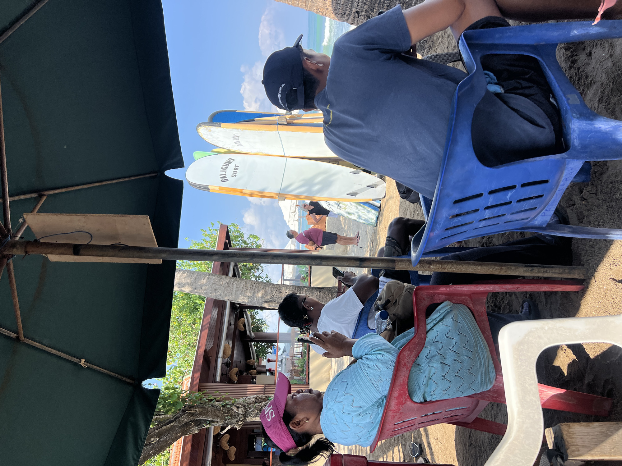 In Kuta, Indonesia, under a large green umbrella on a sandy beach, people sit on chairs. In the foreground, a woman wearing a pink visor and a light blue knitted top is seated in a red chair, holding a mobile phone. Next to her, in a dark chair, a person in a white t-shirt is also looking at their phone. Behind them, several surfboards are propped up. Further in the background, a person in a dark t-shirt and baseball cap faces away from the camera, looking out at the ocean. The sea and sky are visible, with waves breaking on the shore. A building with a thatched roof is situated to the left, with palm trees and lush vegetation nearby. Other individuals are visible in the distance.