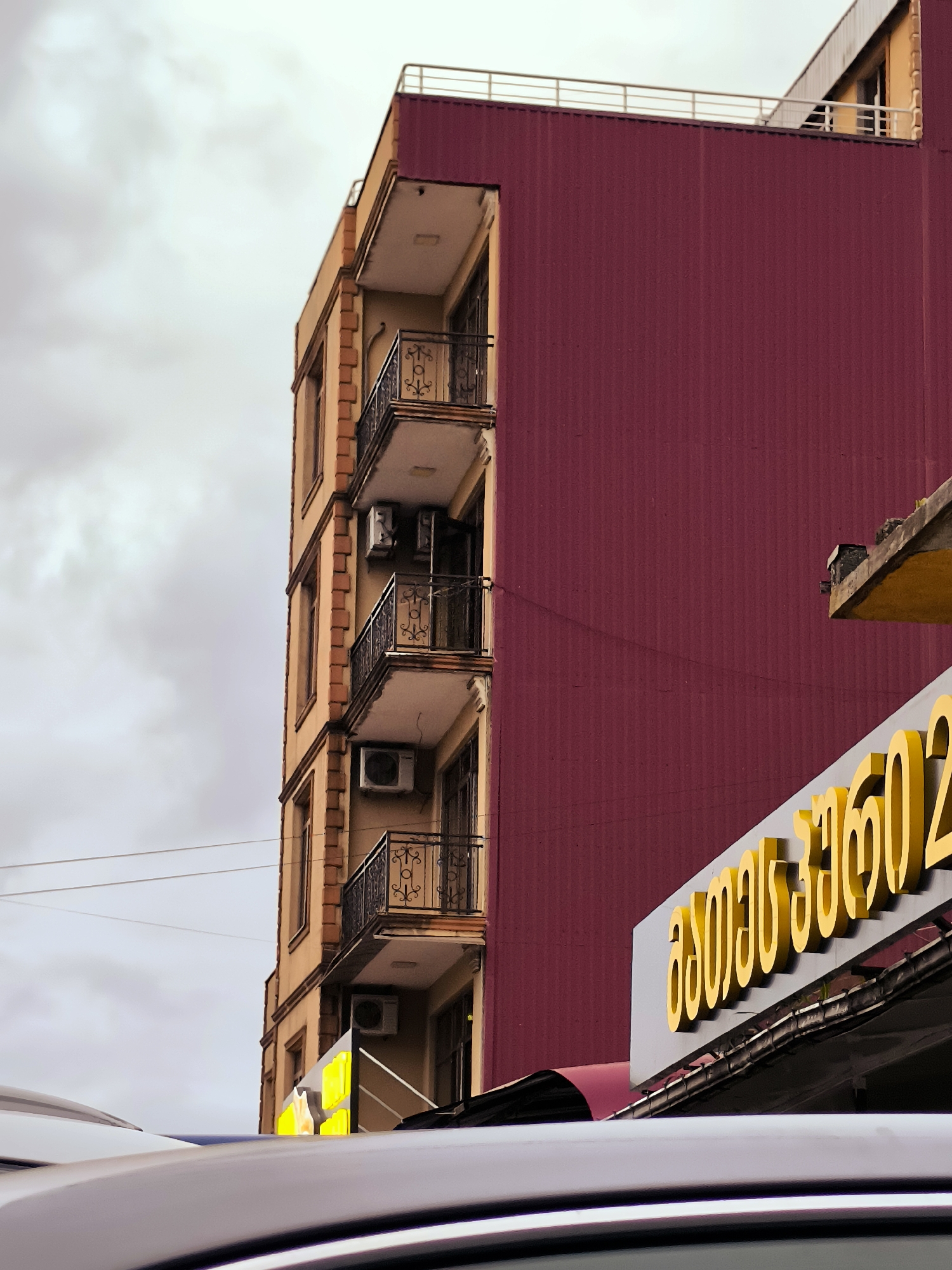The image depicts the side of a tall, narrow building with multiple balconies, likely an apartment building or a mixed-use structure, in Charnali, Georgia. The building has a facade of light brown brickwork and is topped with a dark red corrugated metal section. Several balconies with ornate black metal railings are visible. Air conditioning units are mounted on the exterior walls beneath some of the balconies. In the foreground, the shiny, curved surface of a vehicle's roof is prominent, partially obscuring the lower portion of the building and a yellow sign with Georgian script. The sky above is overcast with clouds.