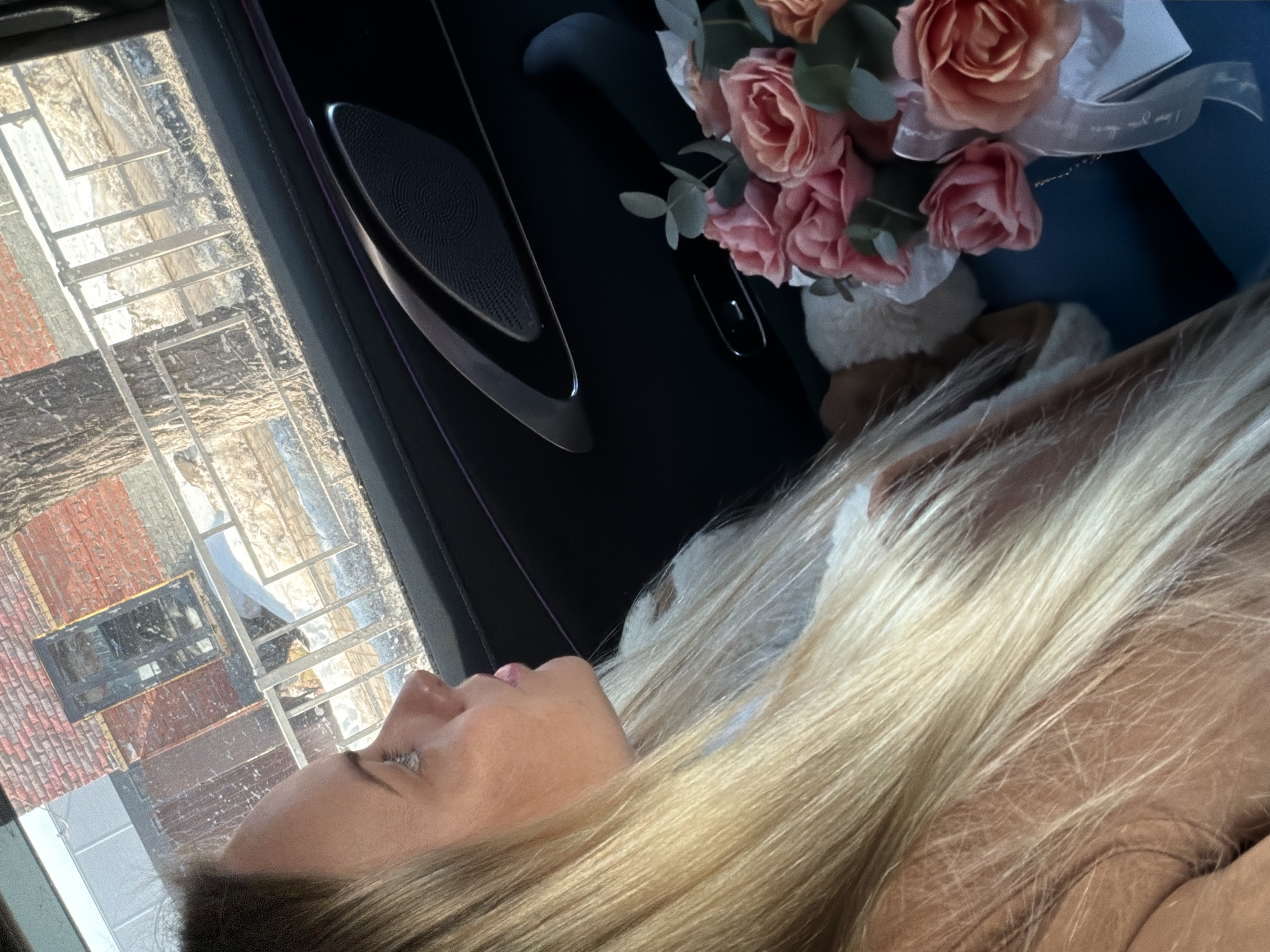 A woman with long blonde hair, seen in profile, occupies the right side of the frame, looking out of a vehicle's window towards the left. Her lips are slightly parted, and her gaze is directed outwards. A bouquet of pink and peach-colored roses, interspersed with eucalyptus leaves and tied with a white ribbon, is situated on a dark blue surface to her right. The interior of the vehicle, specifically a door panel with a speaker grille and a control button, is visible.
Outside the window, a wintry scene in Votkinsk, Russia, is visible. The ground is covered with patches of snow. A metal fence or railing runs parallel to the vehicle, separating the street from the immediate environment. Beyond the fence, a bare-branched tree trunk stands, and further back, a building constructed of red brick with a dark, rectangular window or door is present. The lighting suggests daytime conditions.