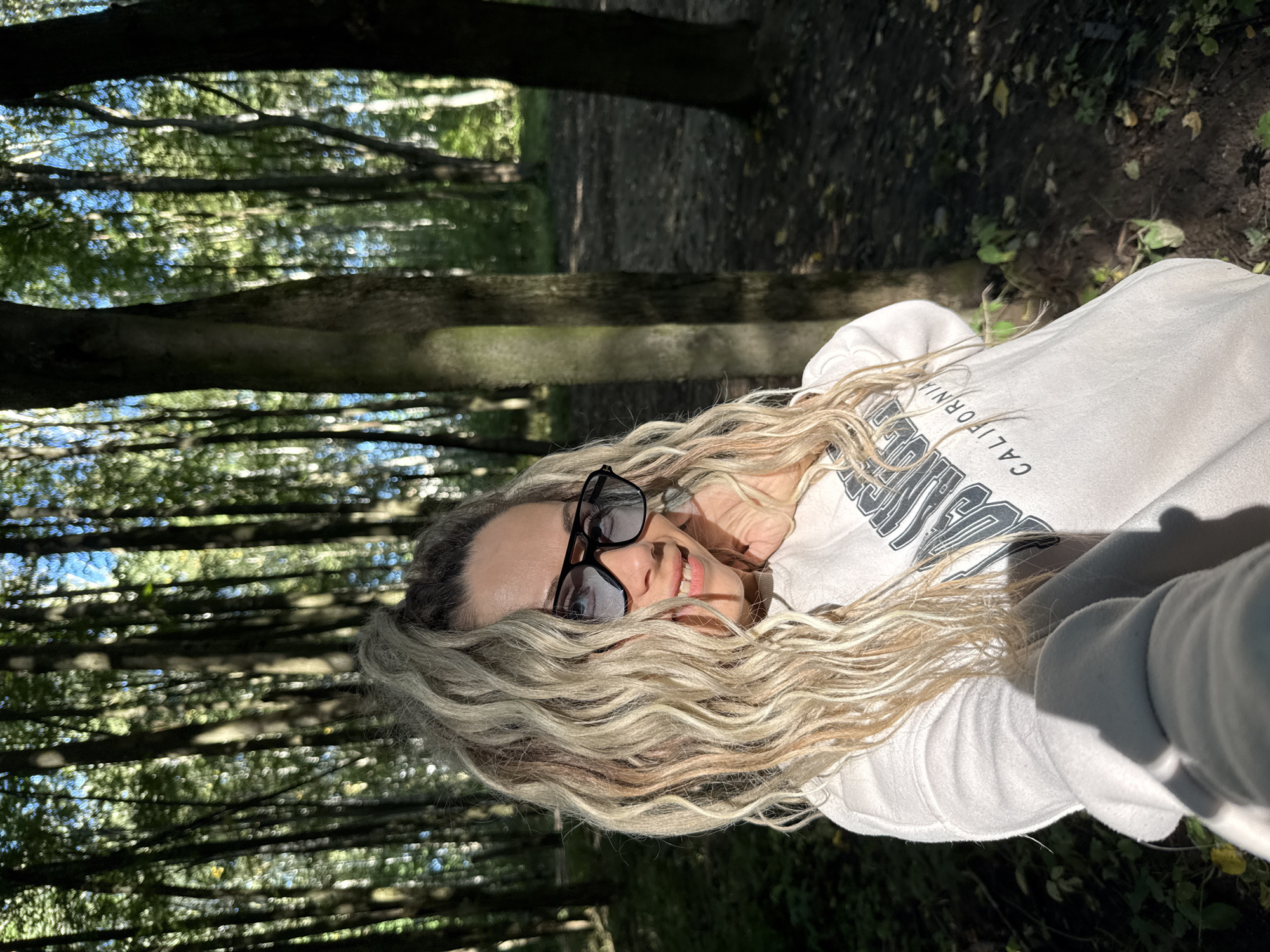 A young woman with blonde dreadlocks is taking a selfie in a forest. She is wearing sunglasses and a white sweatshirt. The woman is smiling and her hand is resting on her chin. The forest is dense with tall trees and sunlight is filtering through the leaves. The ground is covered with fallen leaves. The scene appears to be in the city of Rusanovo, Russia.