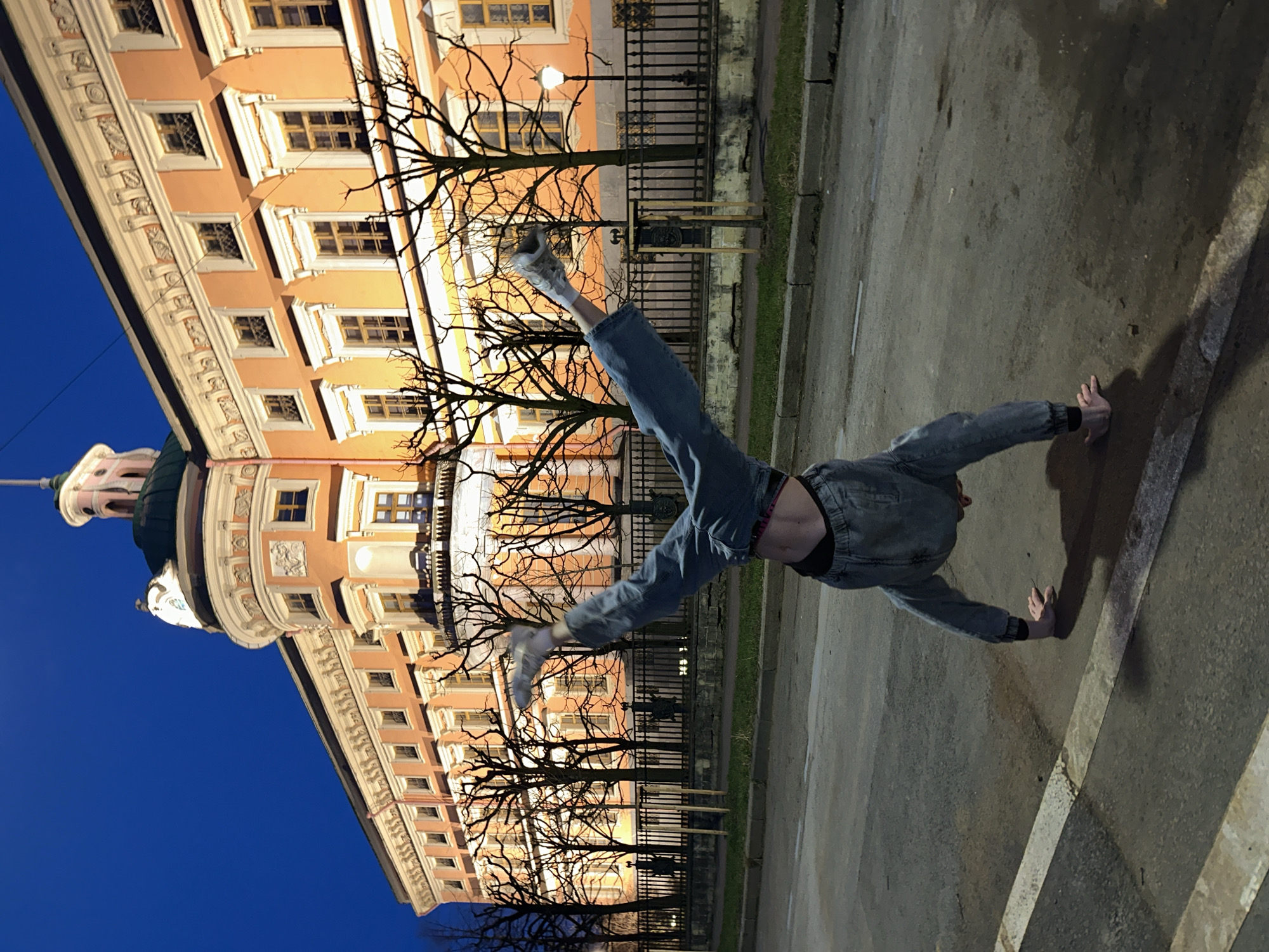 A person is doing a handstand on a paved road with a building in the background. The building is an ornate, multi-story structure with numerous windows, a tower, and decorative architectural details. Bare trees stand between the road and the building, creating a visual layer. The sky above the building is a deep blue, suggesting it's twilight or night. The person is wearing jeans, sneakers, and a top that reveals their midriff. The road is marked with white lines. The location is likely in Saint Petersburg, Russia, given the style of the building.