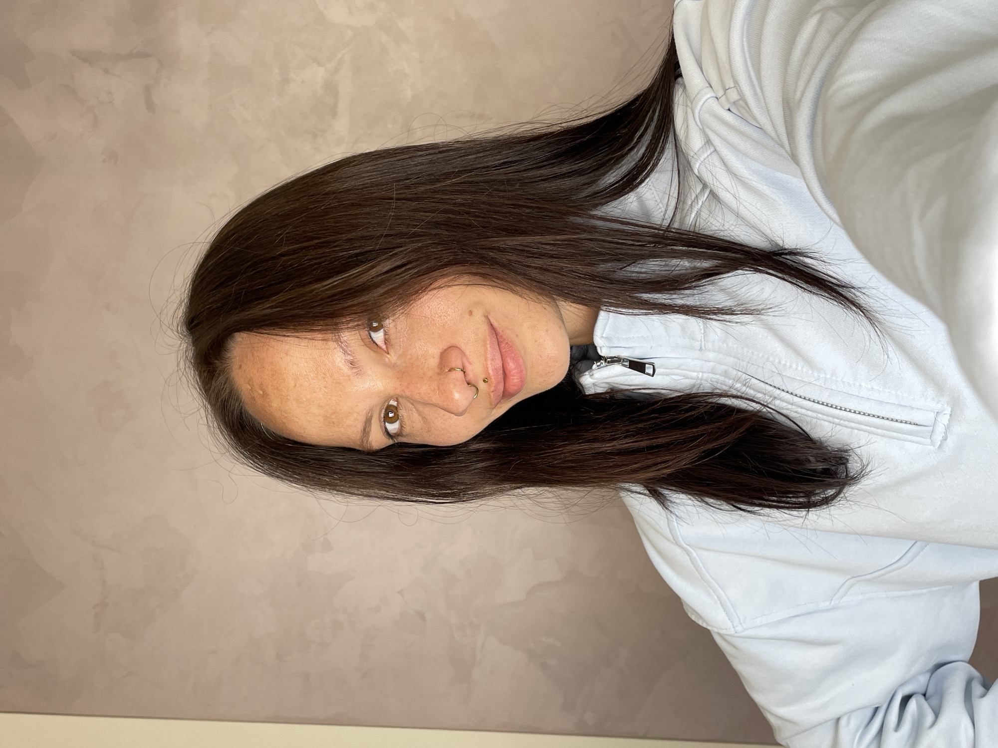A close-up portrait, rotated 90 degrees counter-clockwise, depicts a young woman looking directly at the camera with a subtle, friendly smile. She has long, dark brown hair that flows around her, and warm brown eyes. Freckles are visible across her nose and cheeks. She wears a gold hoop nose ring in her right nostril and a small gold stud piercing above the right side of her upper lip. Her attire consists of a light-colored, possibly light blue or white, zippered top or jacket, with the zipper closure visible near her neck. The background is a plain, textured wall in a light beige or taupe shade, suggesting an indoor setting. There is no visible text. The image was taken in Izhevsk, Russia.