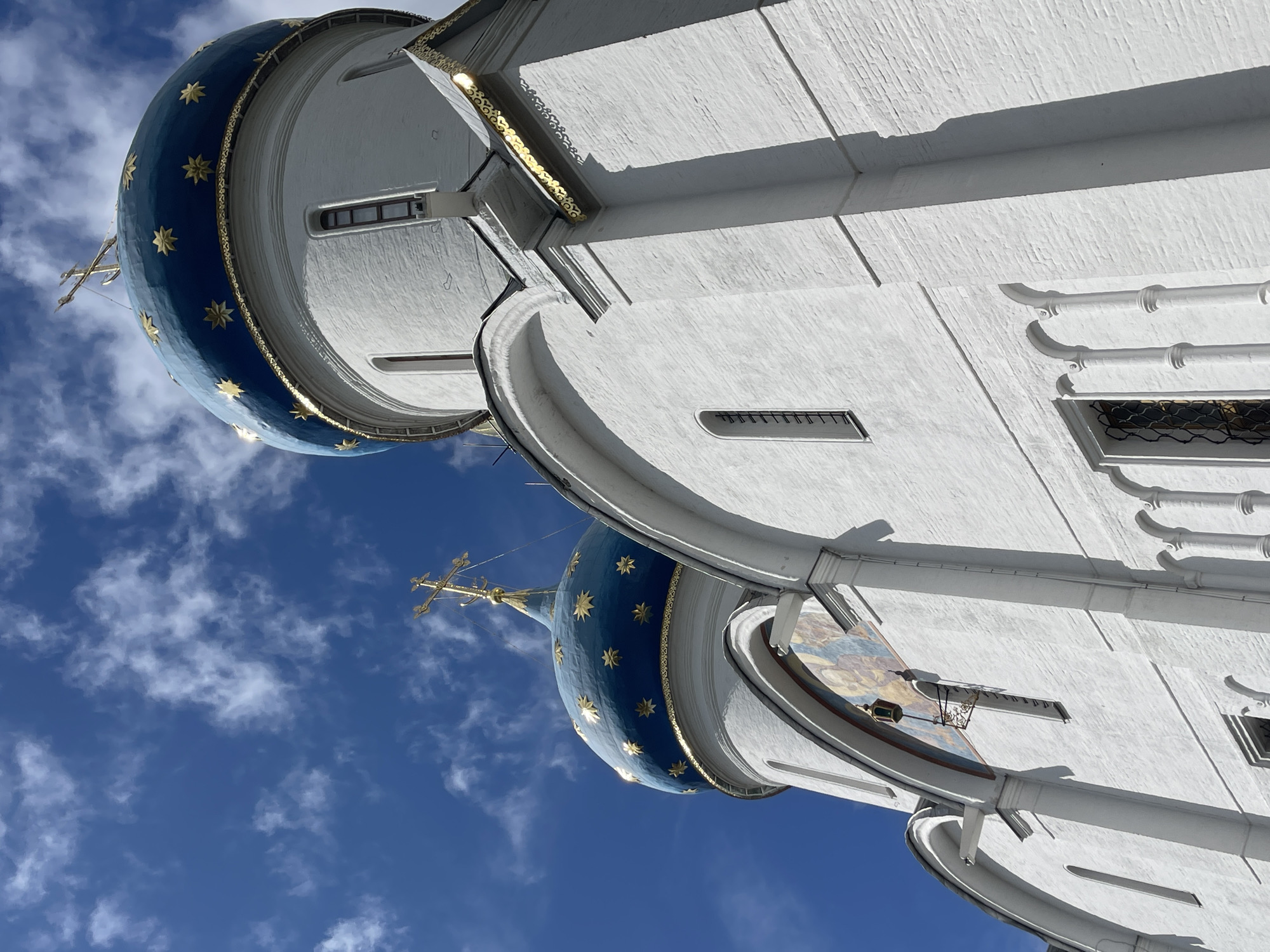This is a low-angle, close-up shot of the white brick exterior of a church with blue domes, located in Sergiyev Posad, Russia. The domes are adorned with gold stars and topped with golden crosses. The sky above is a vibrant blue, scattered with fluffy white clouds. The architecture features arched windows and decorative gold trim along the rooflines. One dome is more prominent in the foreground, while another is visible behind and to the left. The texture of the white brickwork is clearly visible, and the sunlight casts shadows on the building.
